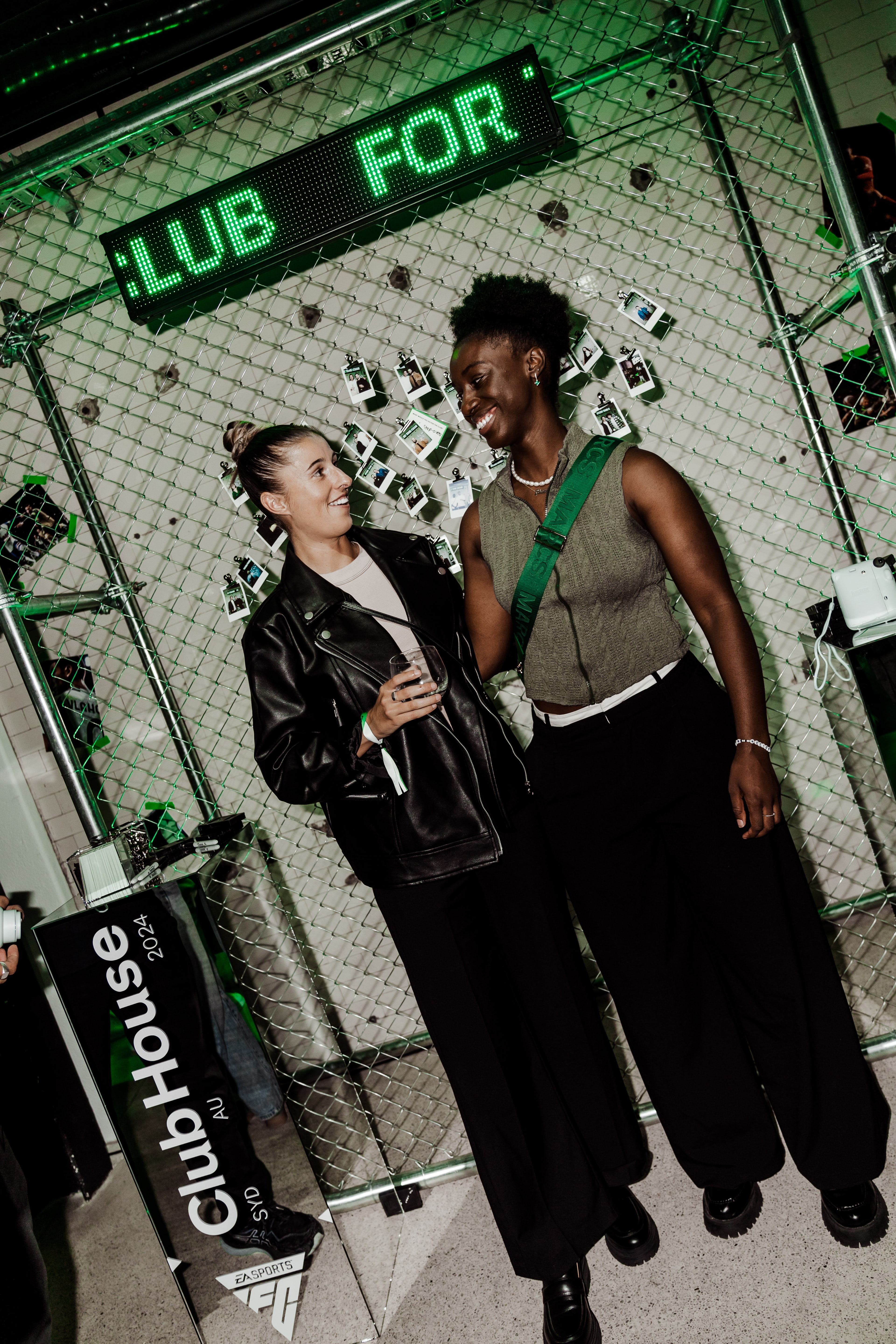 Two smiling women stand together against a wire-fence installation with a green LED ticker reading 'CLUB FOR' beside a ClubHouse plinth.