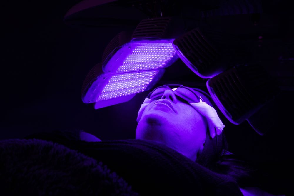 Client in protective goggles receives blue and purple LED light therapy from an overhead panel in a darkened treatment room.