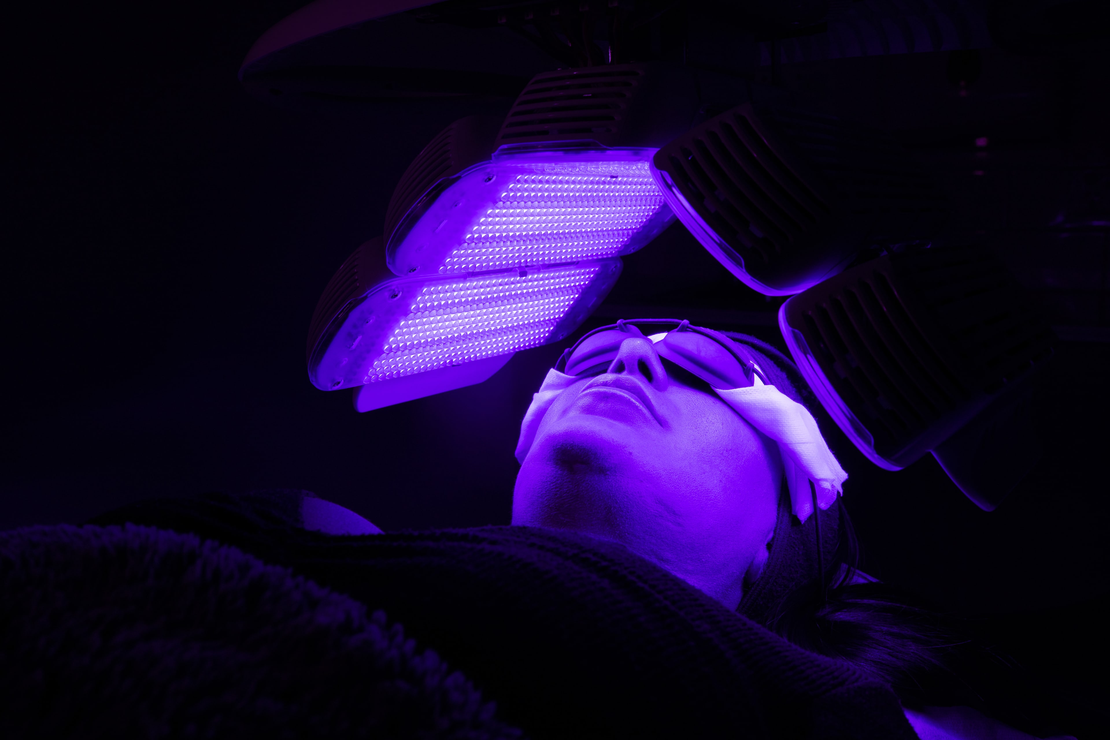 Client in protective goggles receives blue and purple LED light therapy from an overhead panel in a darkened treatment room.