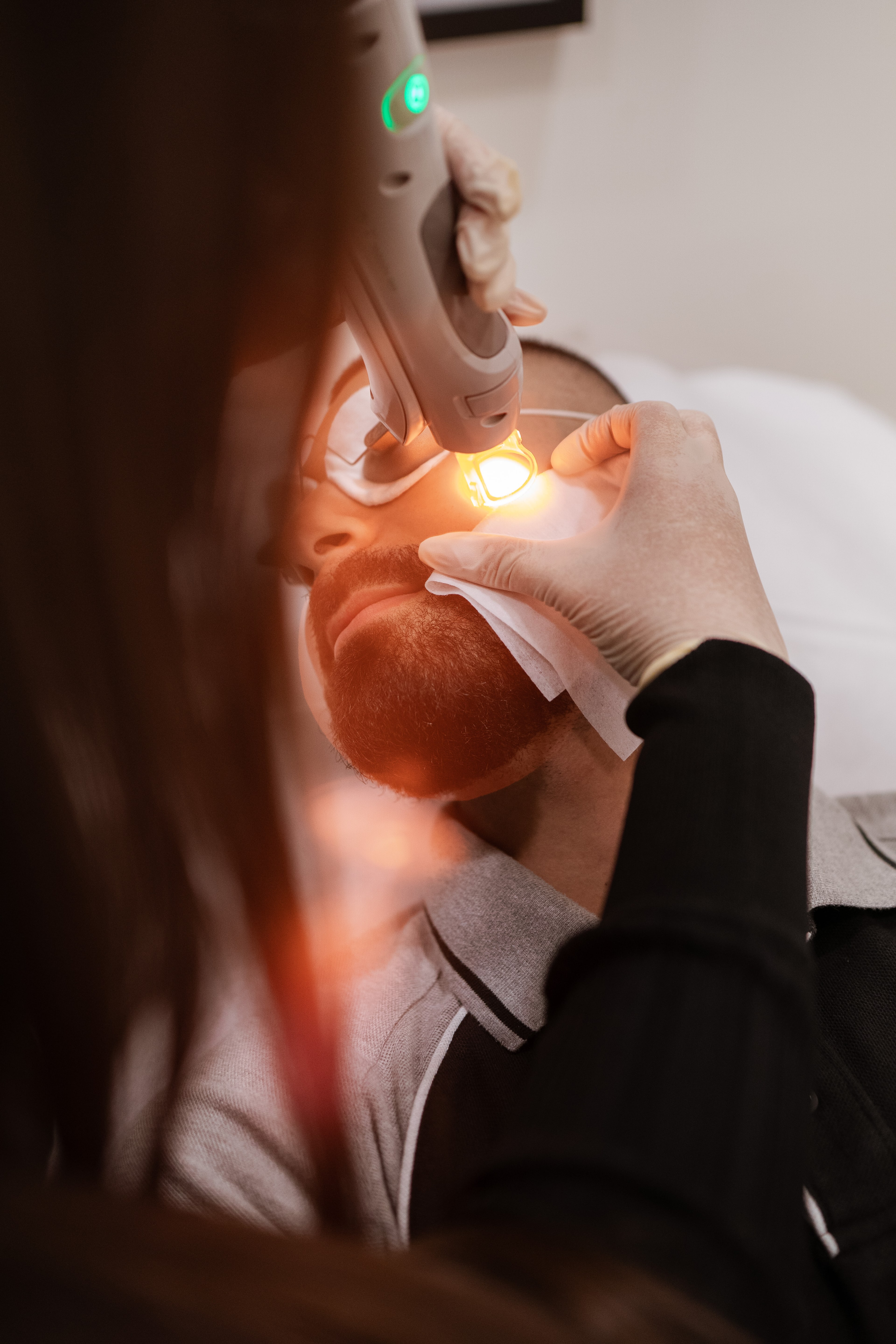 Gloved therapist uses a handheld laser device on a bearded male client's face while holding a gauze pad during skin treatment.