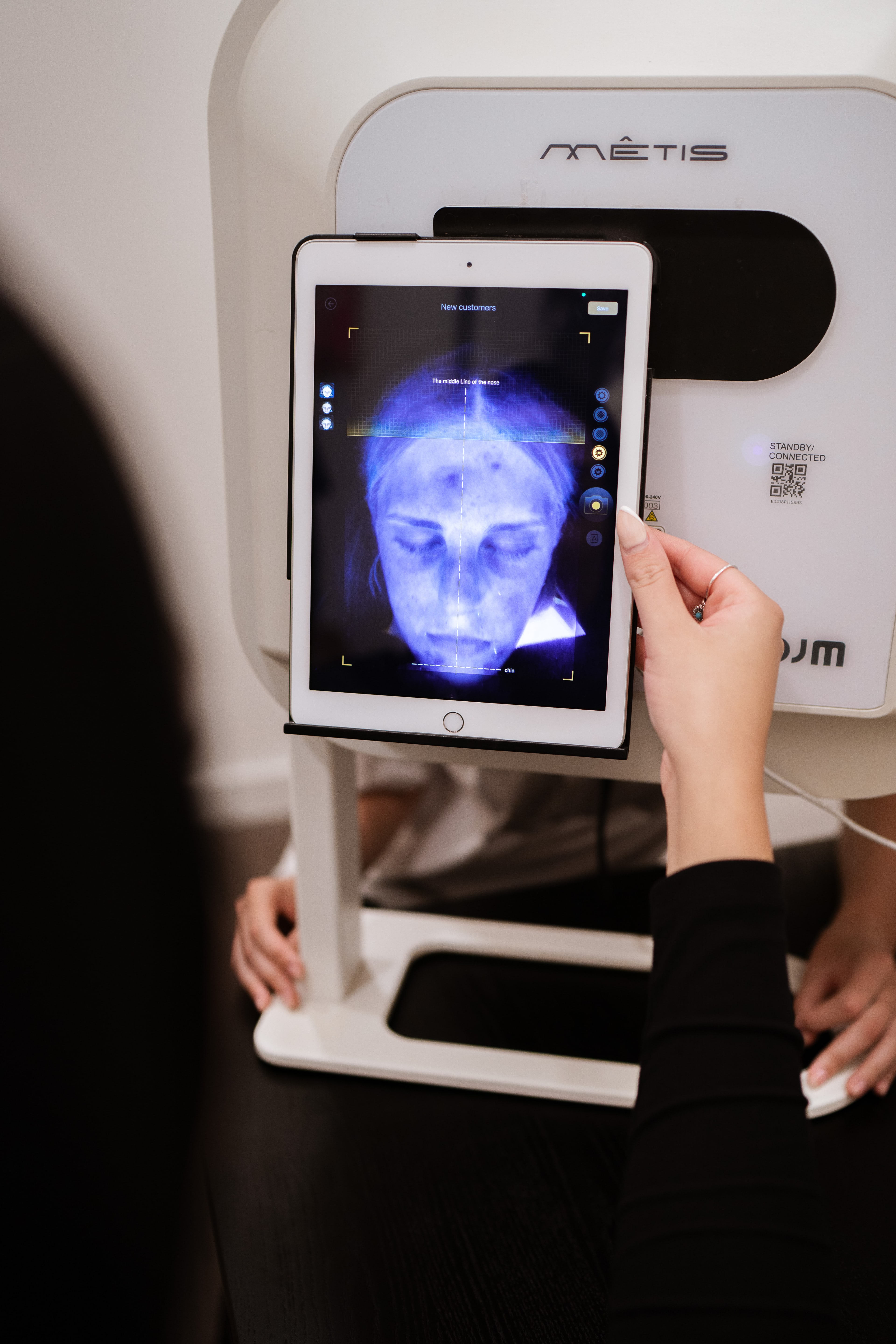 Therapist holds a tablet on a Metis skin-analysis machine displaying a UV facial scan of a client during a consultation.