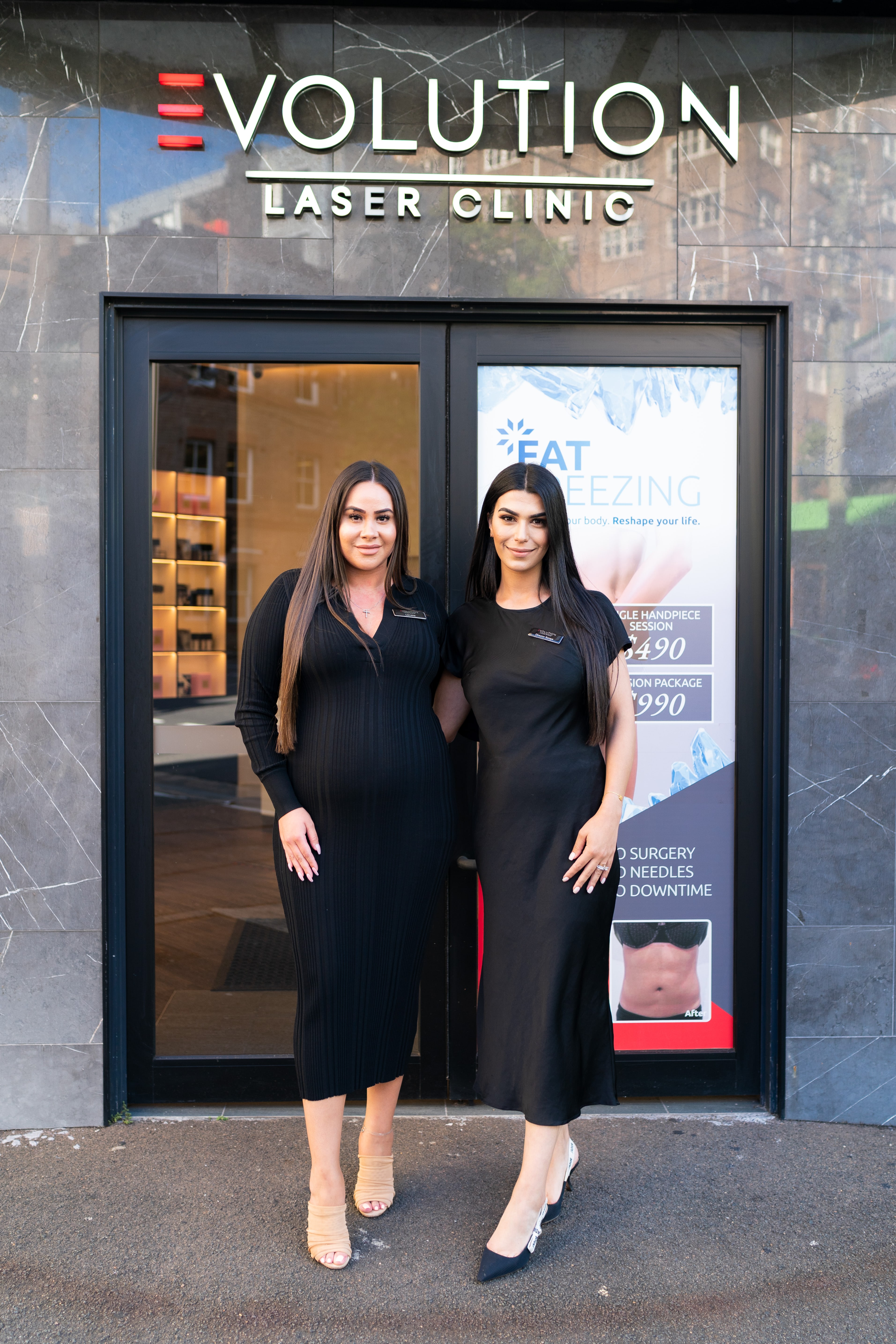 Two women in black dresses stand smiling outside the Evolution Laser Clinic entrance beneath its signage and a fat-freezing promotional poster.