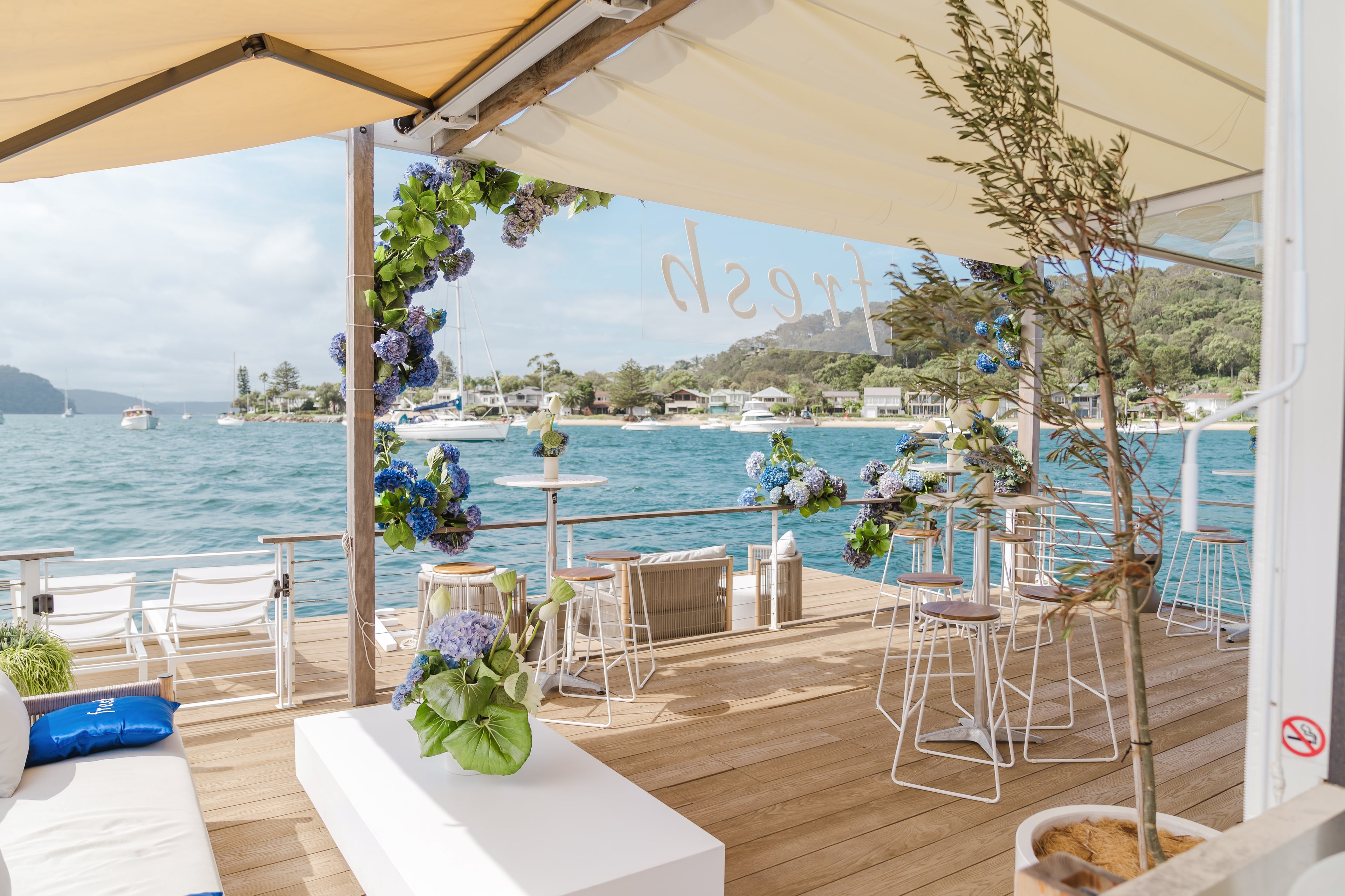Waterfront event deck with blue-and-white hydrangea floral arch, white bar stools and lounge seating overlooking Pittwater at the Fresh launch.