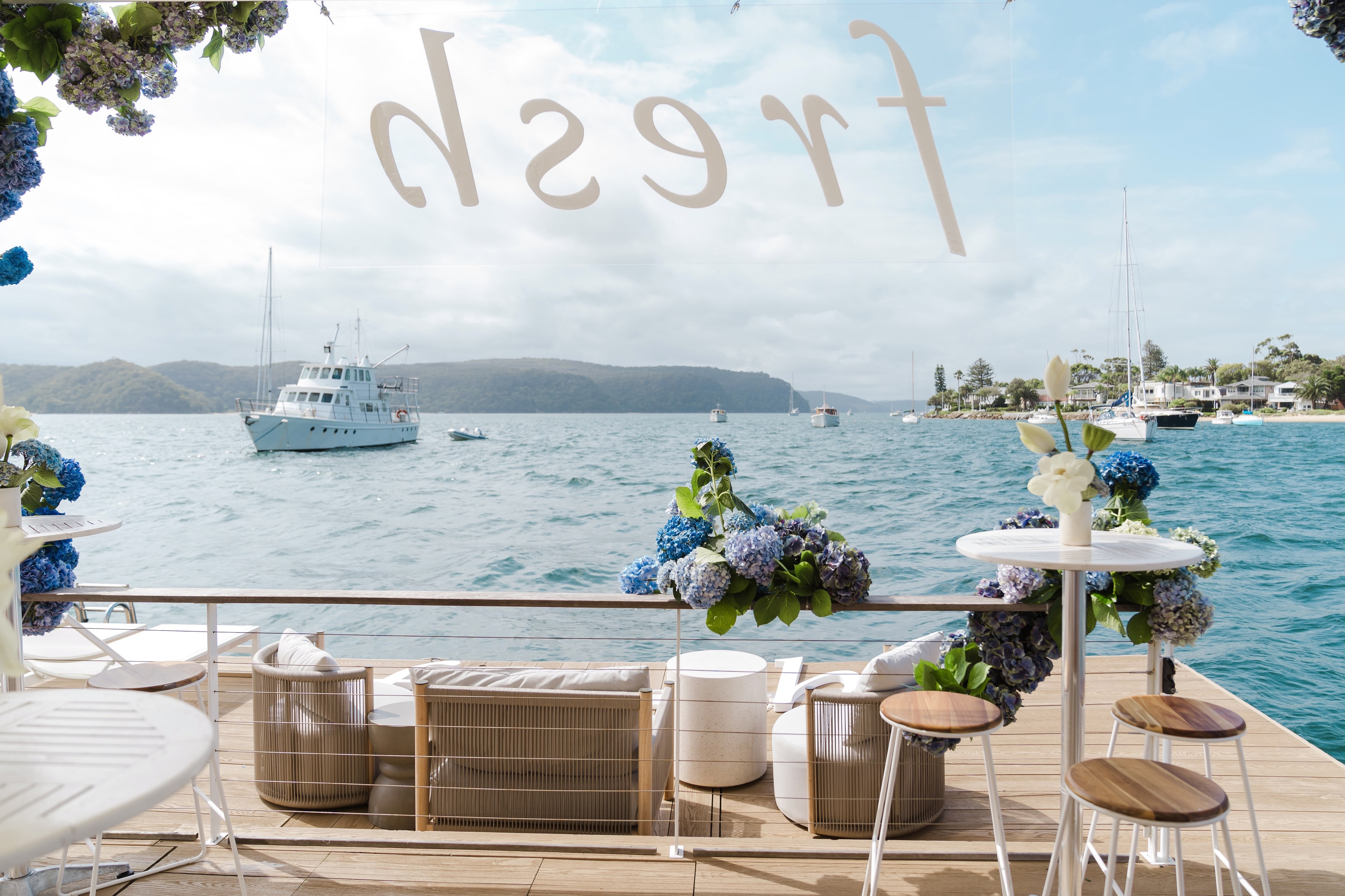 View from the deck through a window etched with the Fresh logo, moored boats on the bay framed by hydrangea-topped cocktail tables.
