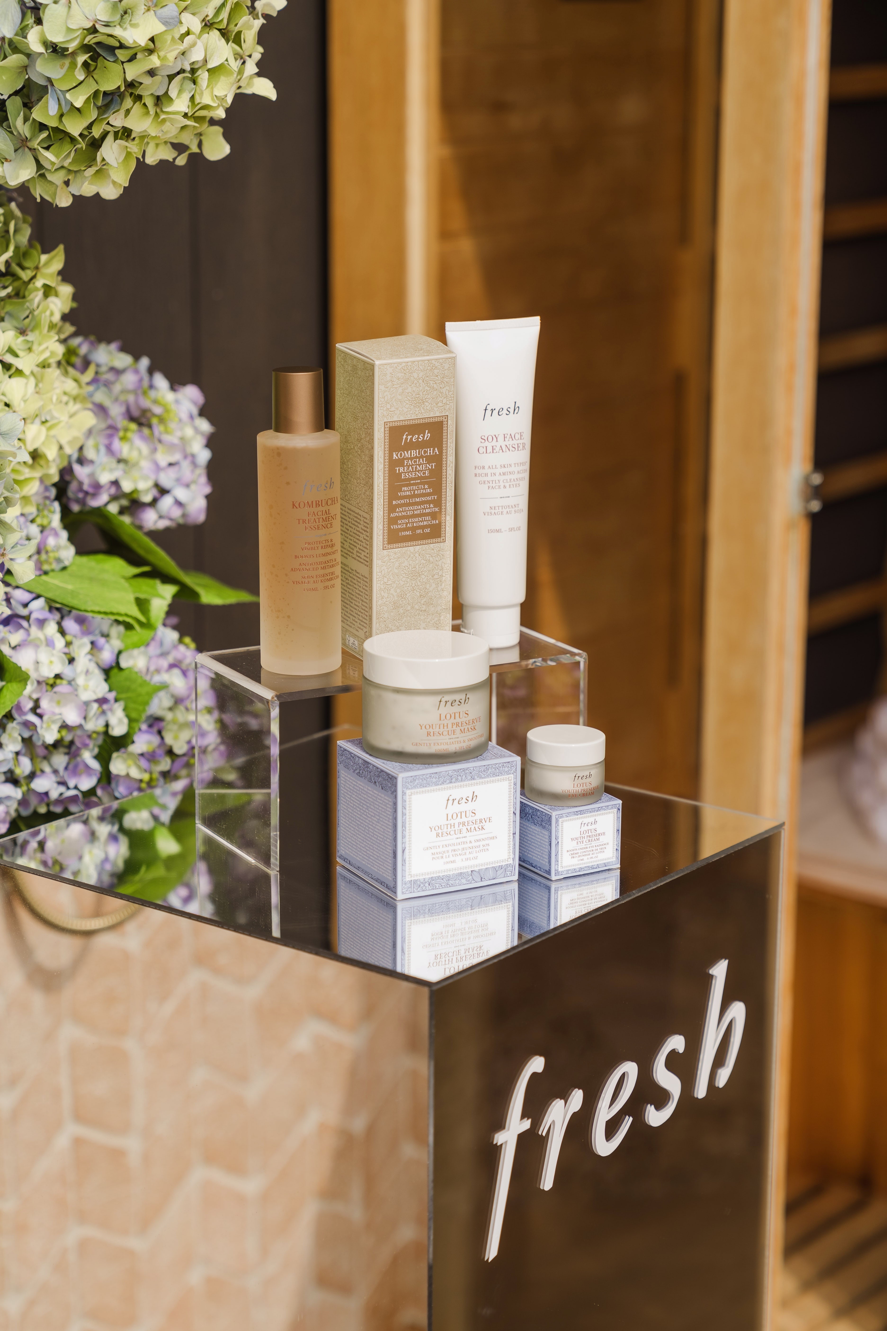 Fresh Lotus skincare range displayed on a mirrored plinth with the Fresh logo, framed by green and purple hydrangeas.
