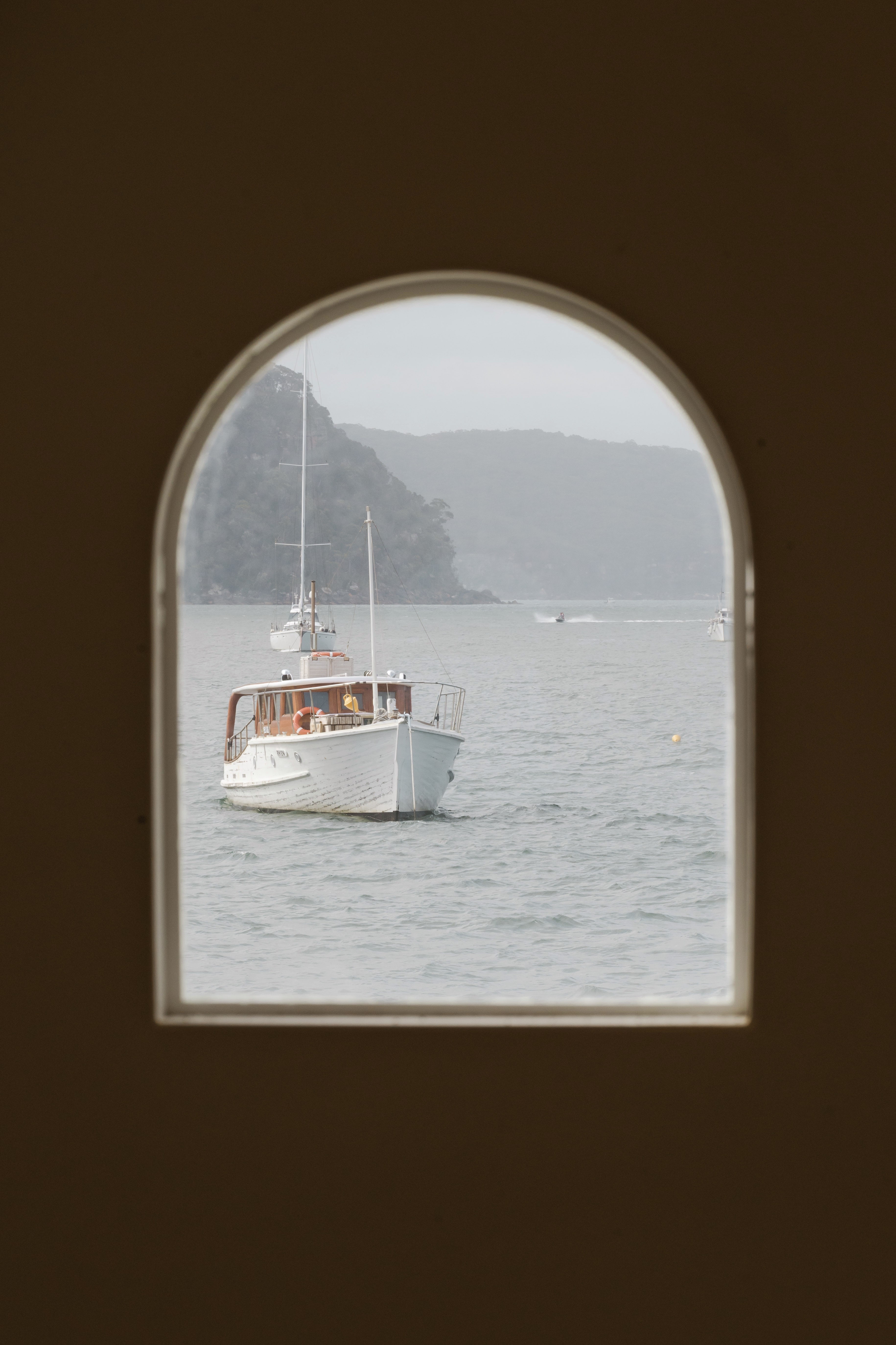 A classic white timber boat moored on the bay, seen through an arched window with misty headland behind.