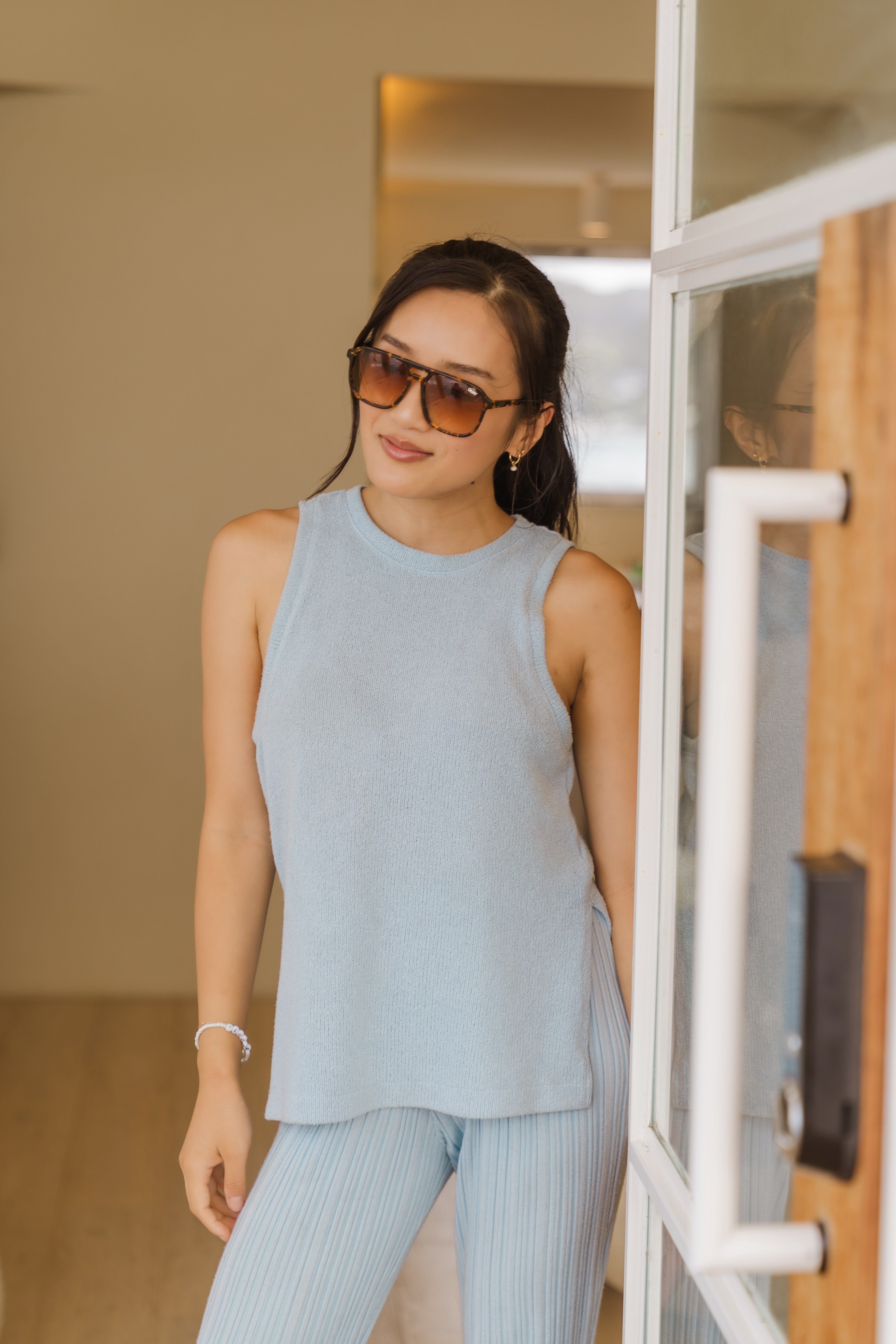 Guest in a pale blue knit tank and matching pleated pants wearing tortoiseshell sunglasses, leaning against a glazed doorway.