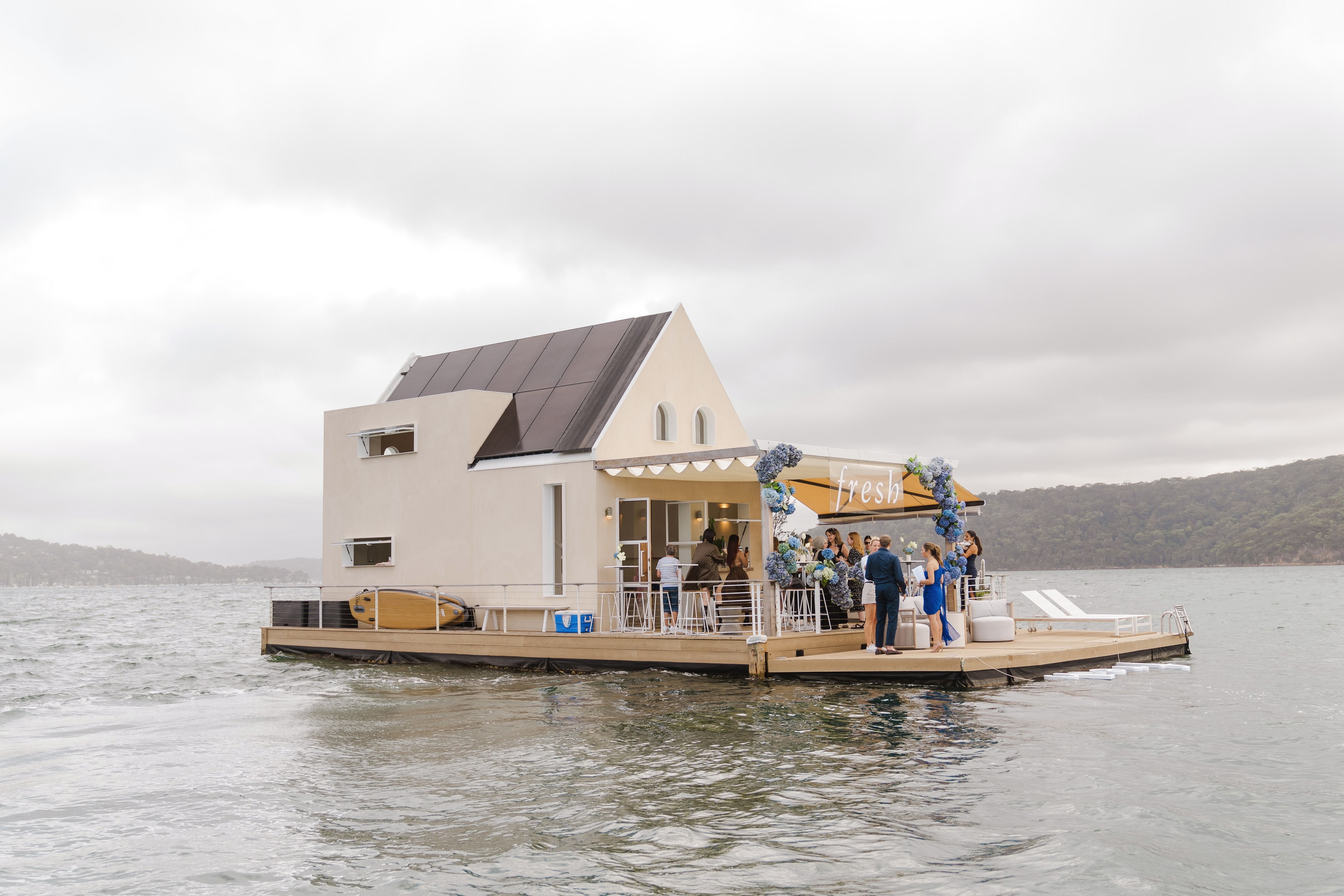 The floating Fresh pavilion on Pittwater under overcast skies, guests gathered on its deck beside a blue floral archway.
