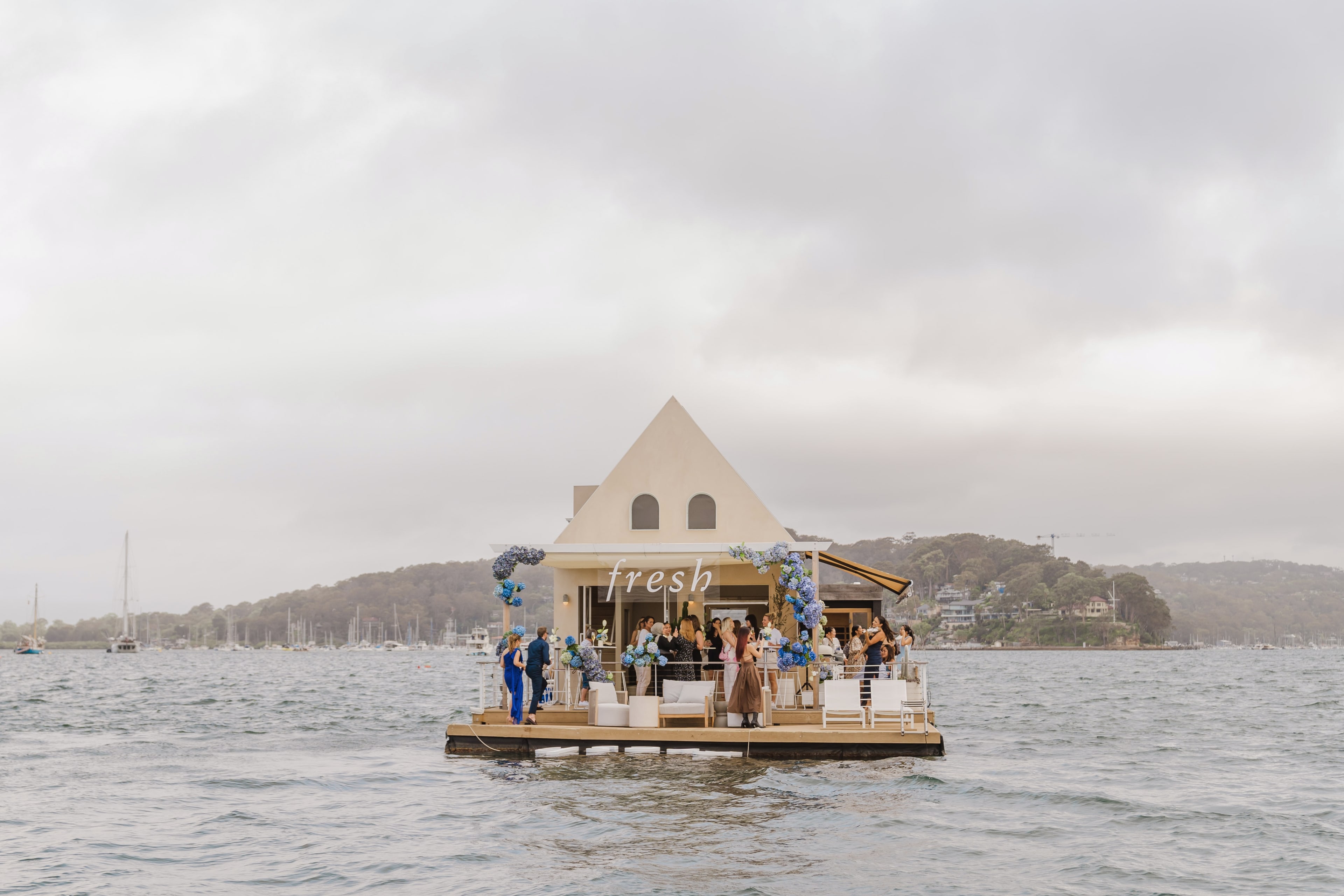 Wide view of the floating Fresh cottage on the bay, guests mingling on the deck between blue hydrangea arches and boats beyond.