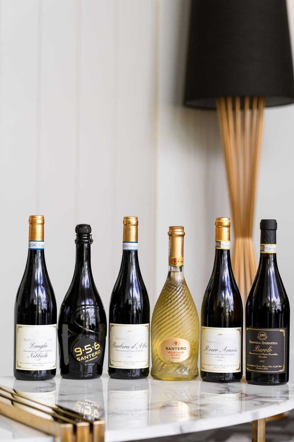 Six Italian wine bottles, including Santero and Barbaresco, lined up on a marble bar cart beside a black-shaded floor lamp.