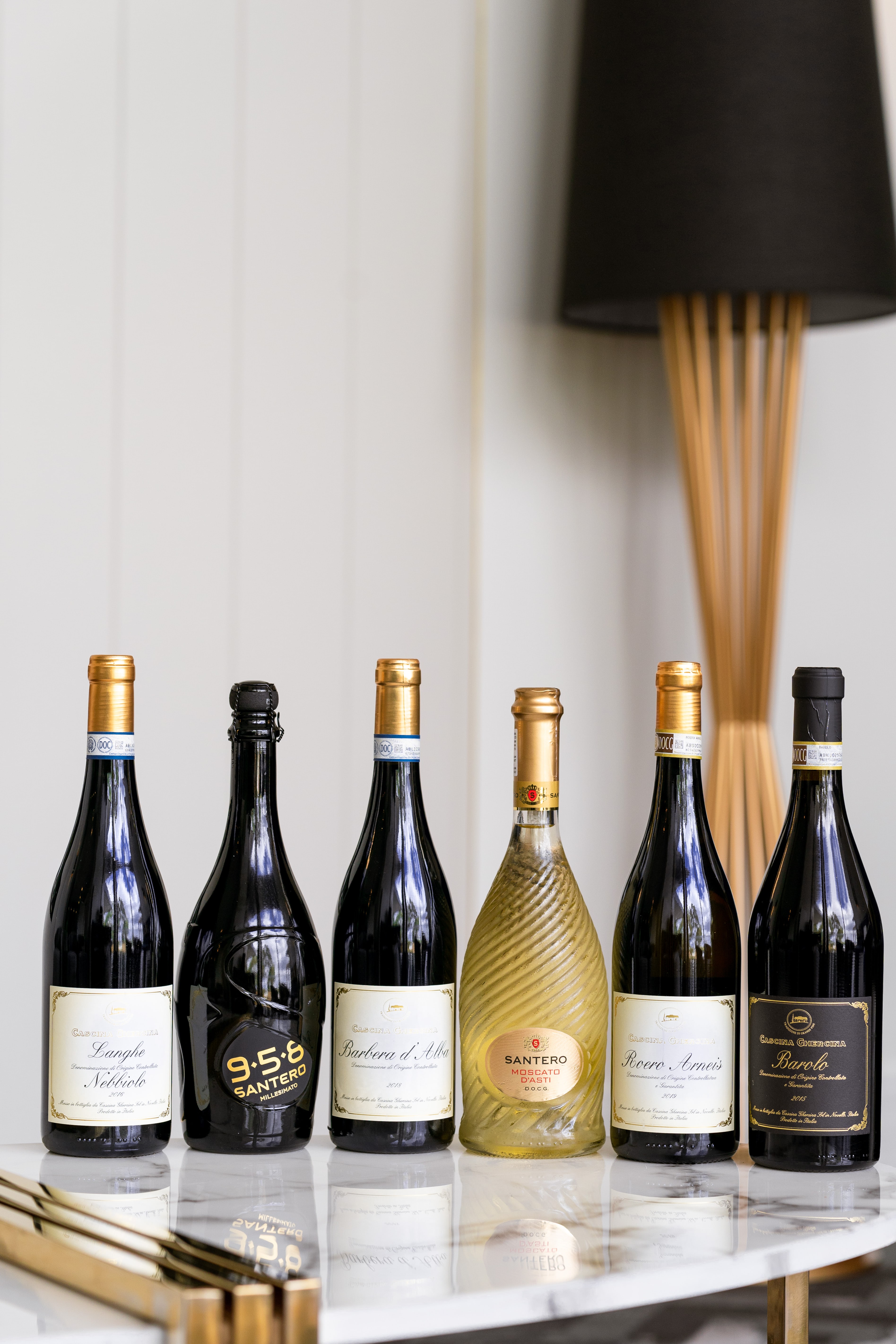 Six Italian wine bottles, including Santero and Barbaresco, lined up on a marble bar cart beside a black-shaded floor lamp.
