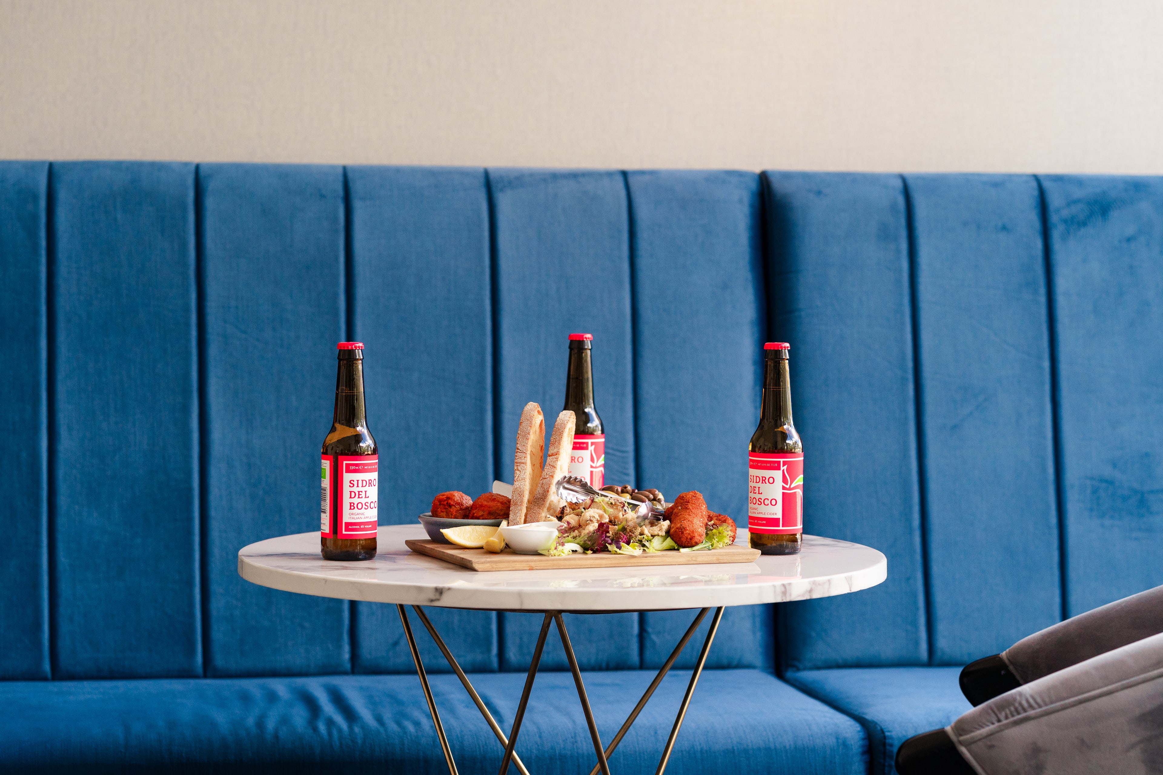 Three red-labelled Reino del Rosco beer bottles on a round marble table with a shared antipasto board, against a blue velvet banquette.