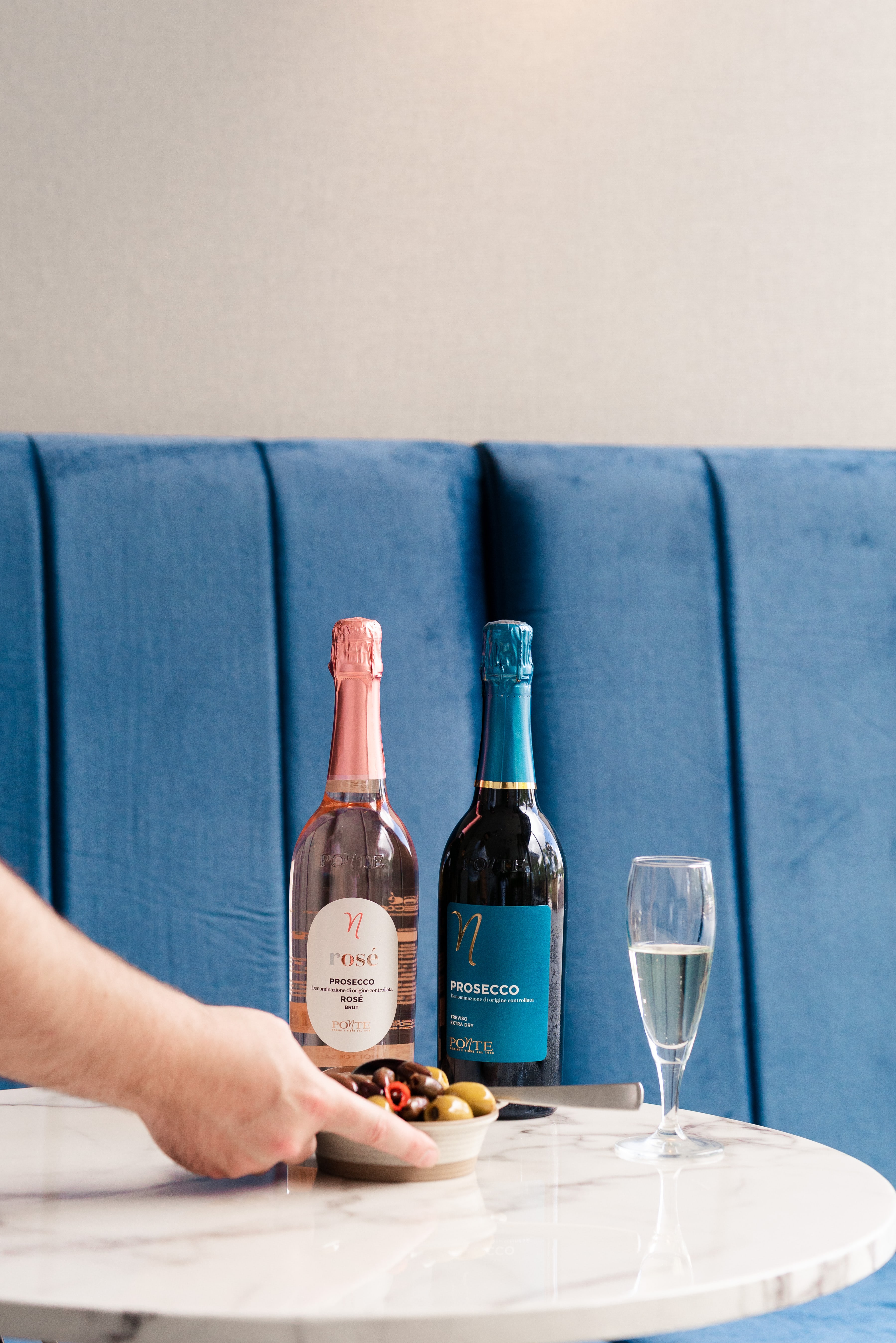 A hand reaches for a bowl of olives beside Ponte rose and Prosecco bottles and a filled flute on a marble table by a blue banquette.
