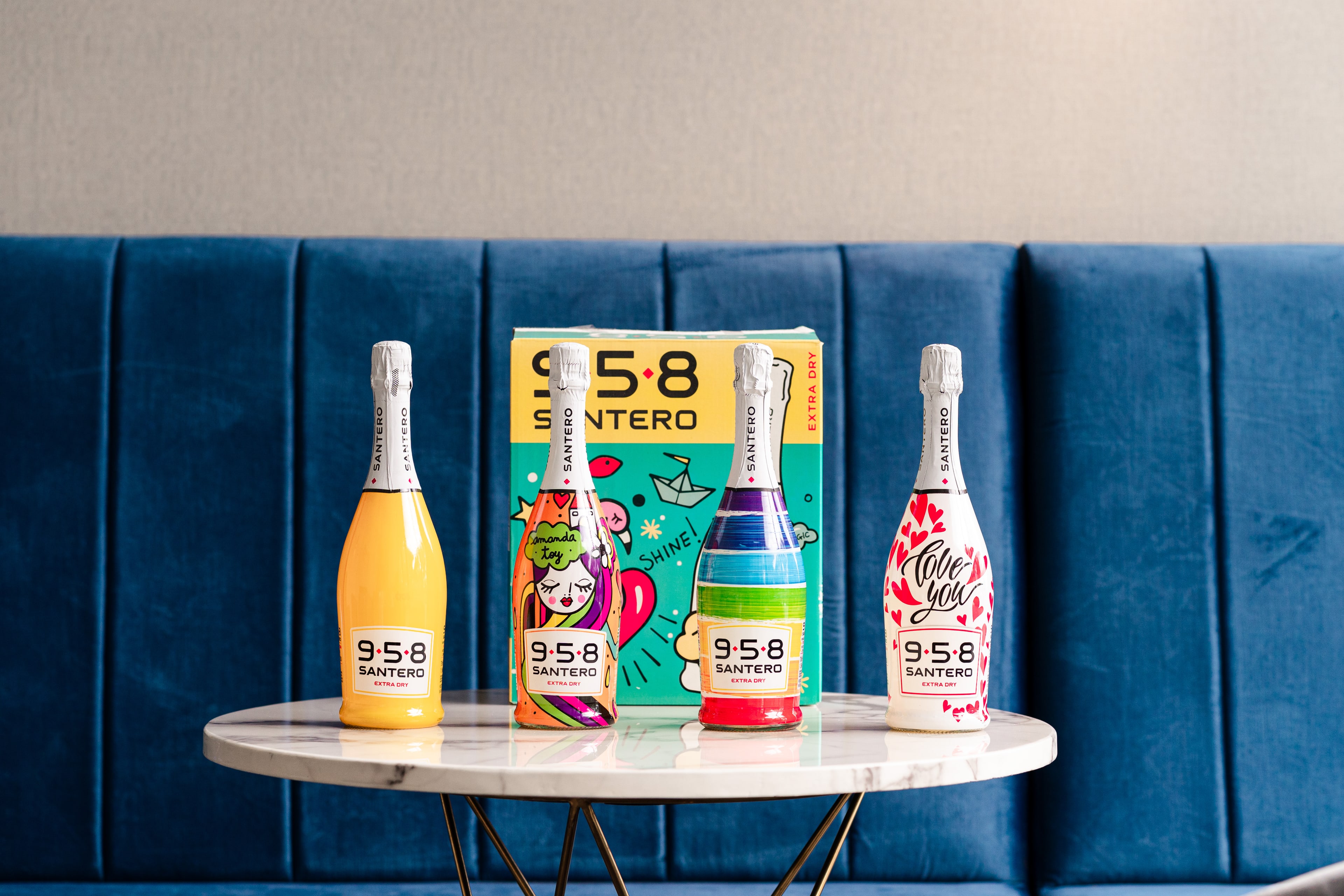 Two colourful Santero 958 sparkling wine bottles flank a printed gift box on a marble table against a blue velvet banquette.