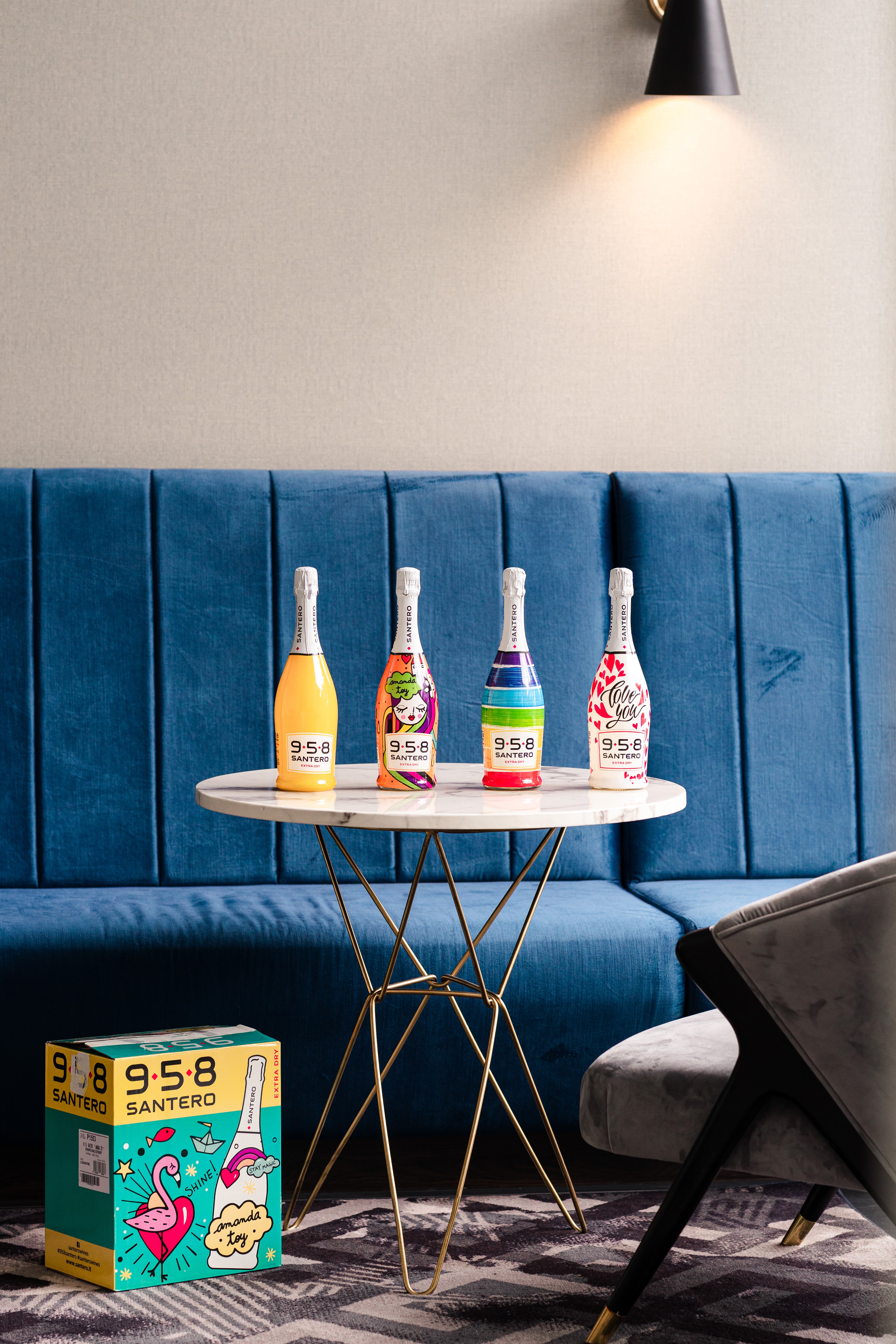 Four vividly decorated Santero 958 sparkling bottles on a marble table, with a matching flamingo gift box on the floor by a blue banquette.