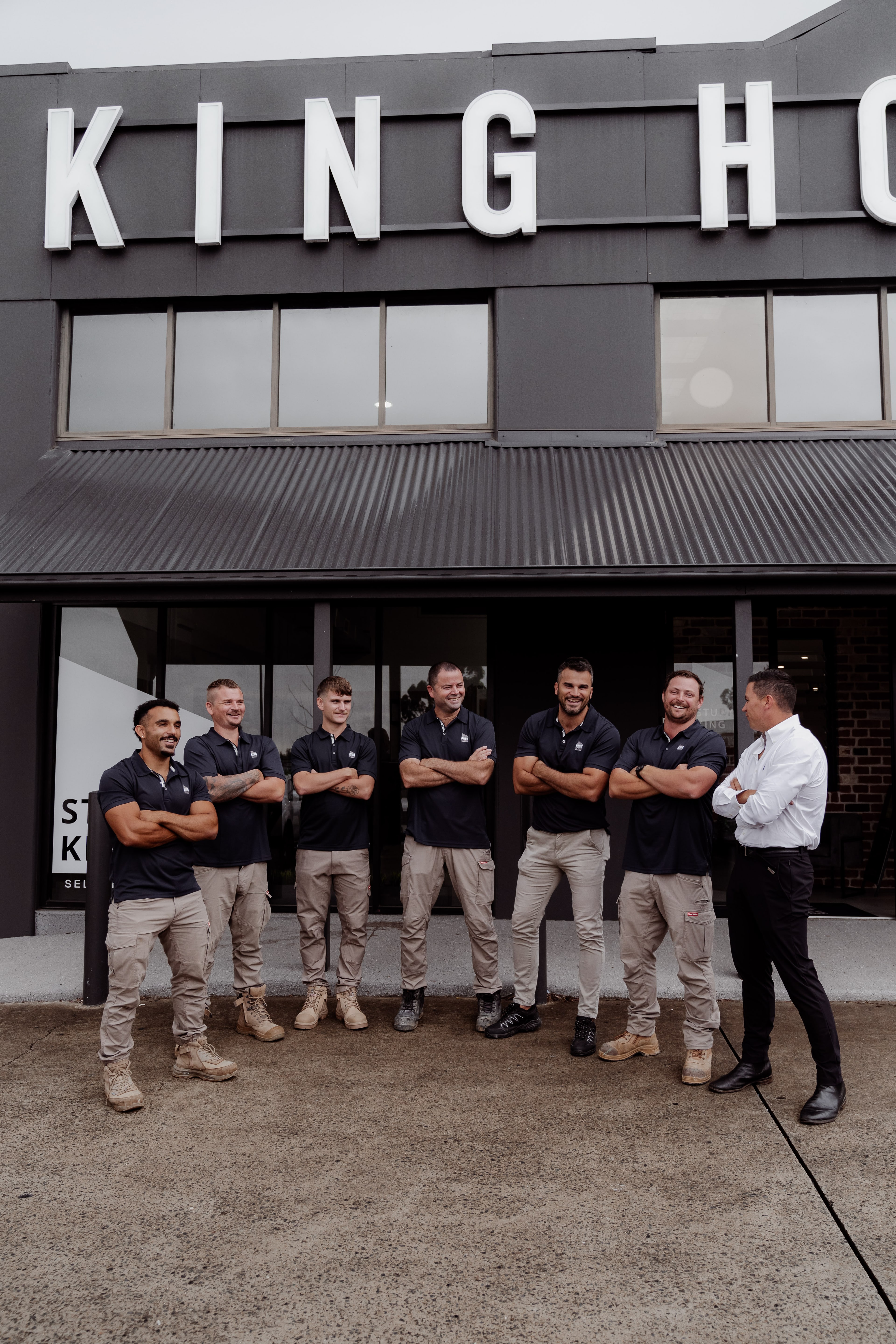 Six King Homes tradesmen in navy polos and chinos stand arms-crossed outside the office as a manager in a white shirt talks with them.