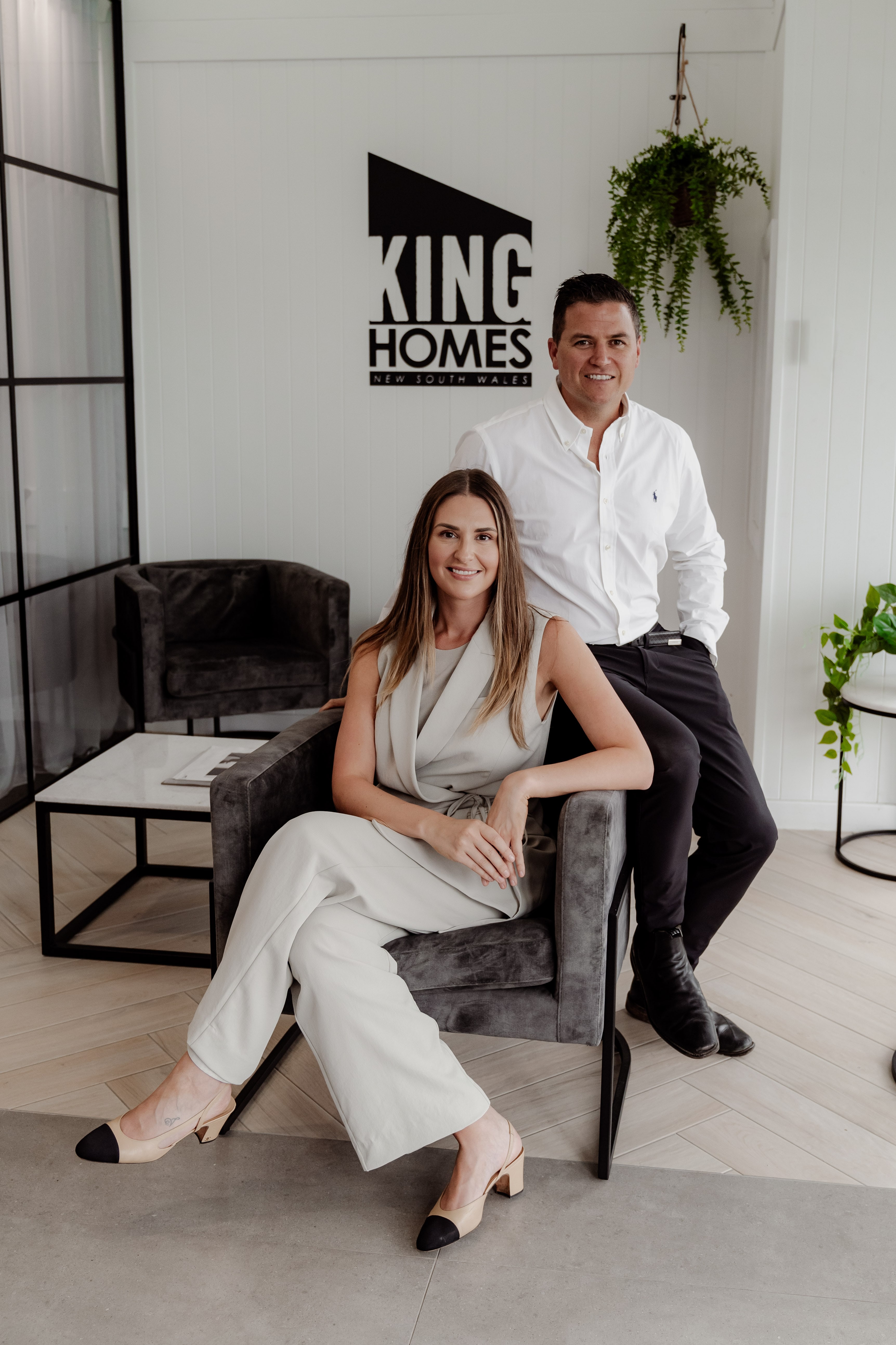 A woman in a cream suit sits in a grey armchair beside a man in a white shirt, in front of the black King Homes NSW wall logo.