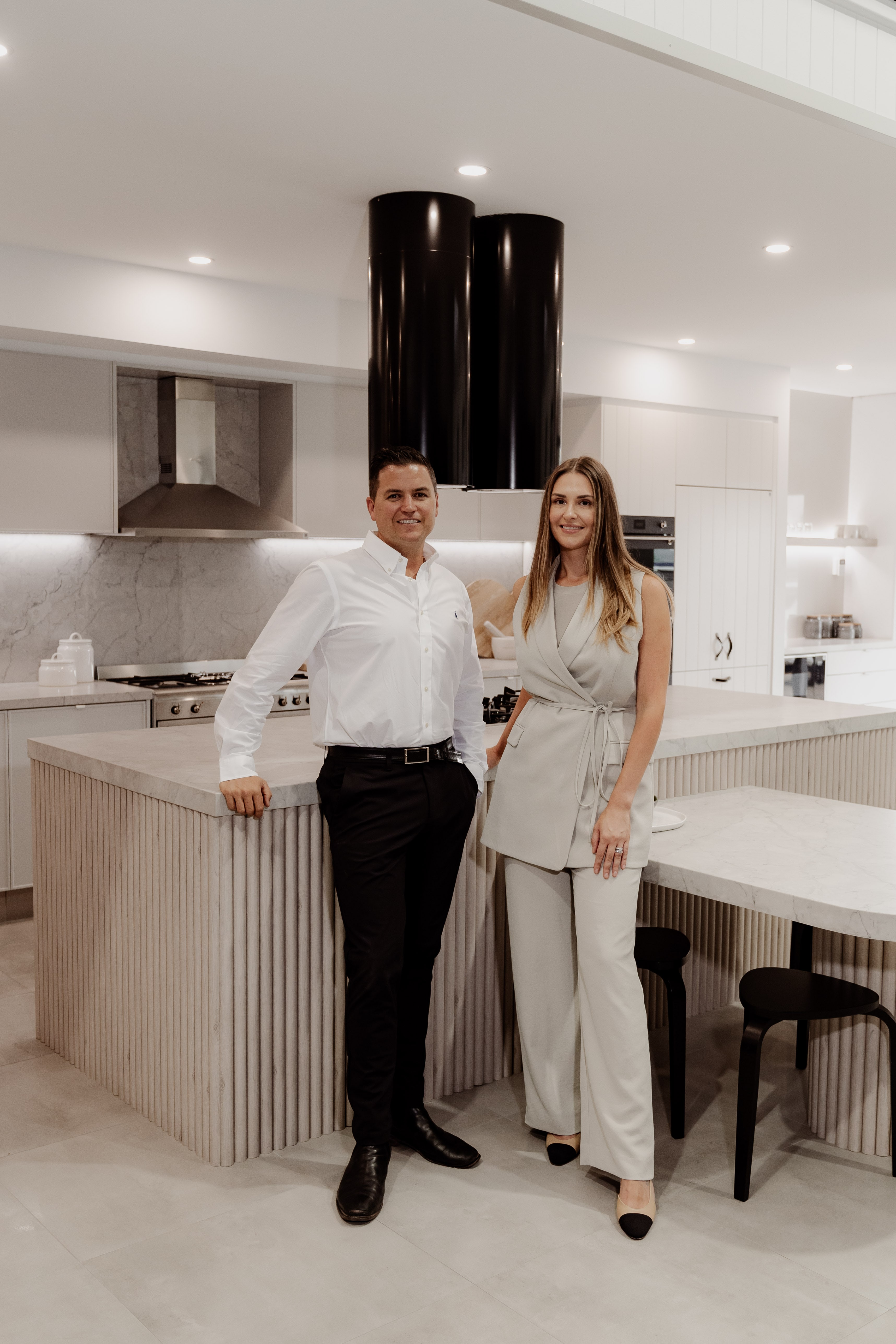 A man and woman in smart attire stand smiling in a display-home kitchen with fluted island benches, marble tops and black cylinder rangehoods.