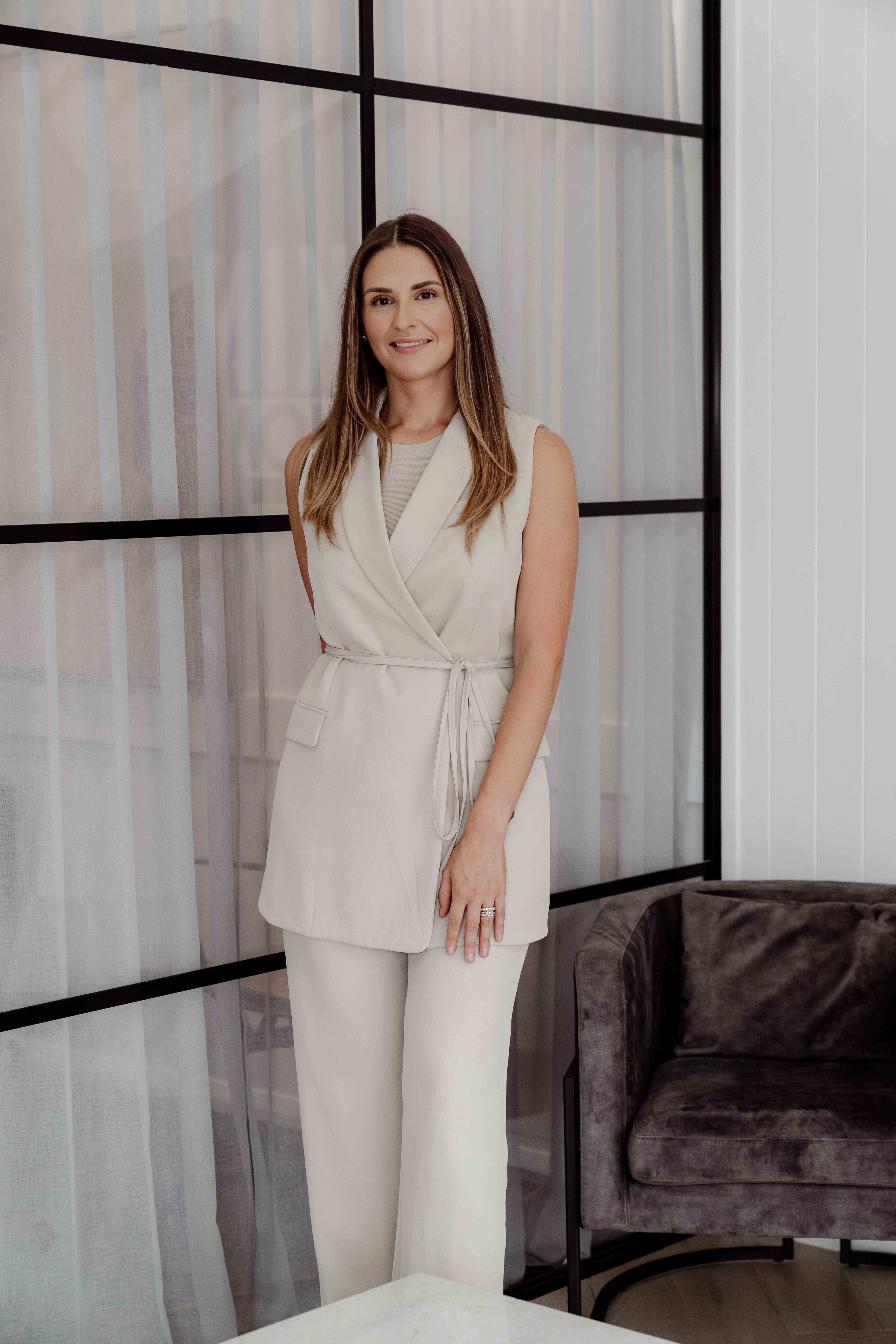 A woman in a cream sleeveless suit stands smiling beside a black-framed sheer glass partition and grey velvet armchair in a bright interior.