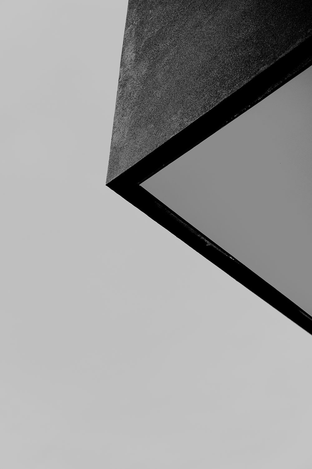 Abstract close-up of a matte black angular roof eave and its slim reflective glazed soffit against a plain grey sky.