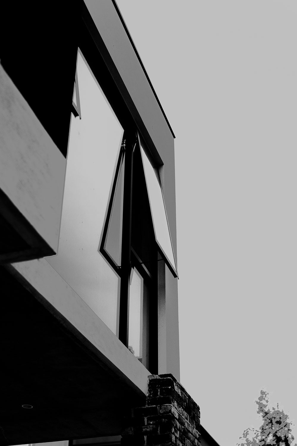 Black-and-white upward view of an awning window opening outward beneath a cantilevered eave, with a brick pier and tree against the sky.