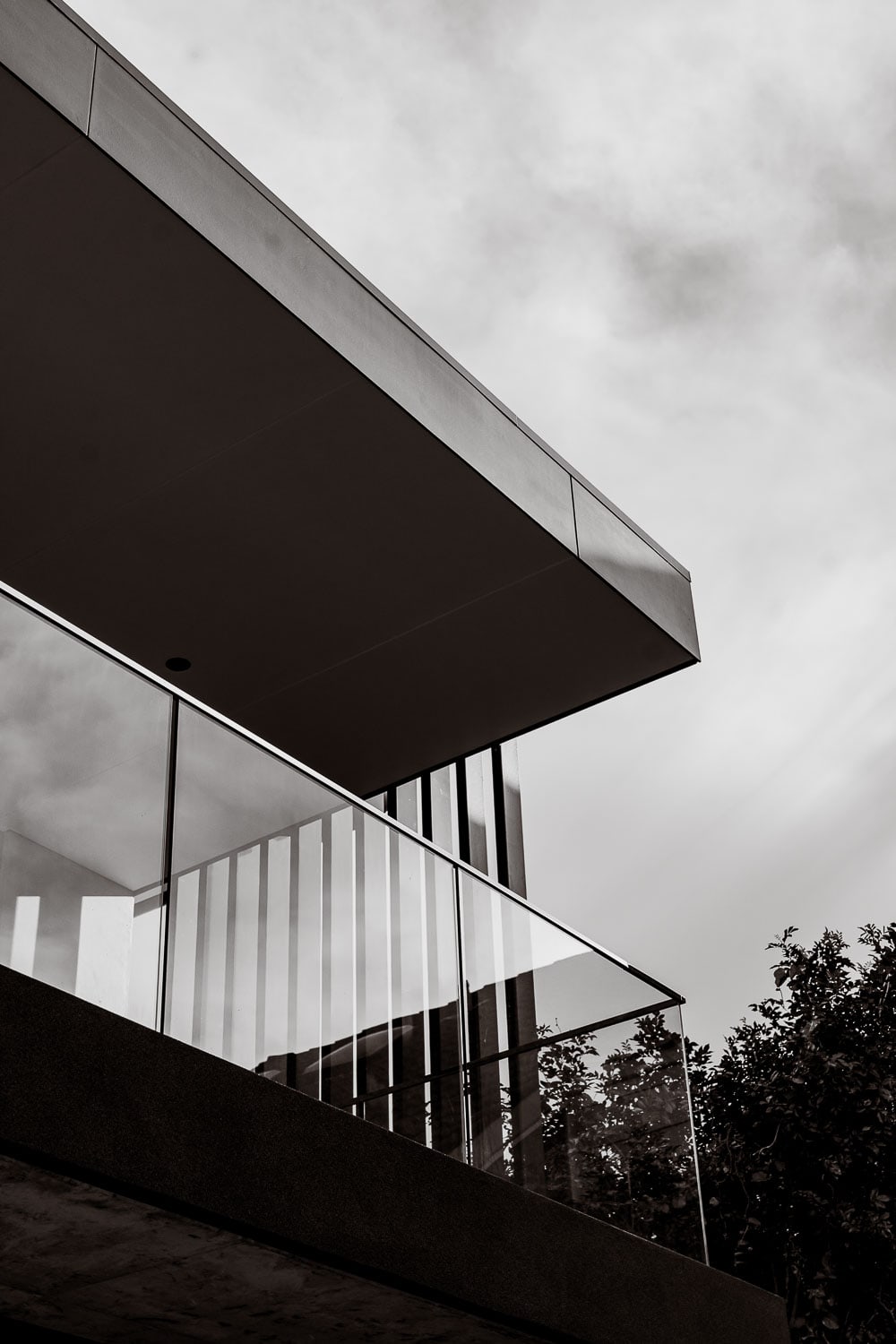 Cantilevered upper storey with a glass balustrade balcony and vertical screen, seen from below against a cloudy sky with foliage at right.