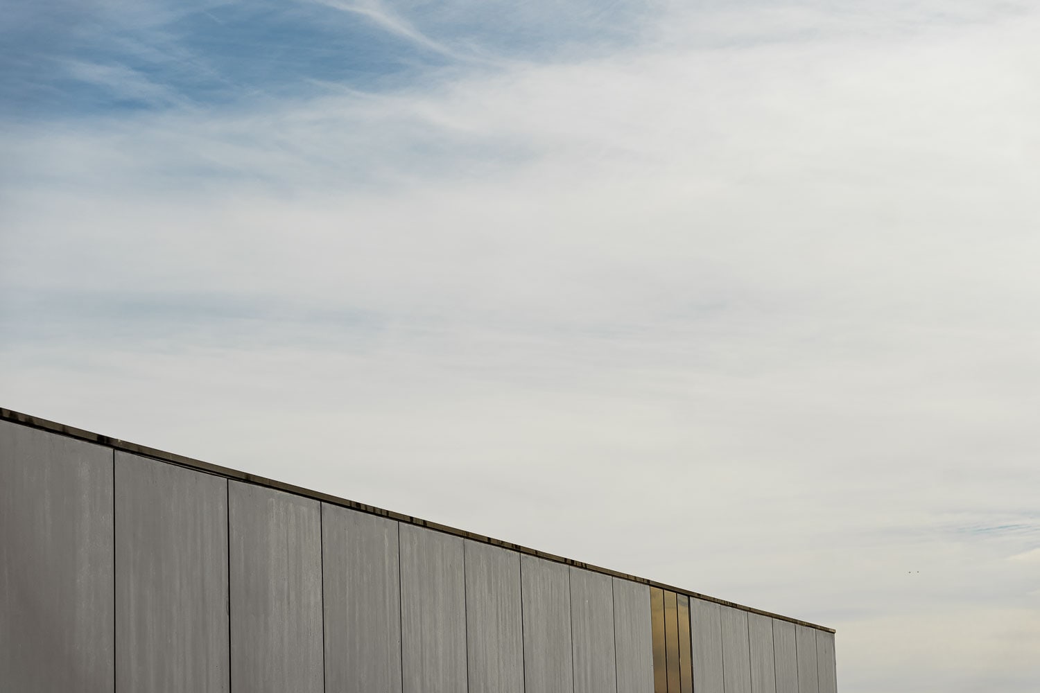 Grey vertical-panelled concrete wall angling up against a blue sky with wispy cloud, a slim brass reveal breaking the facade.