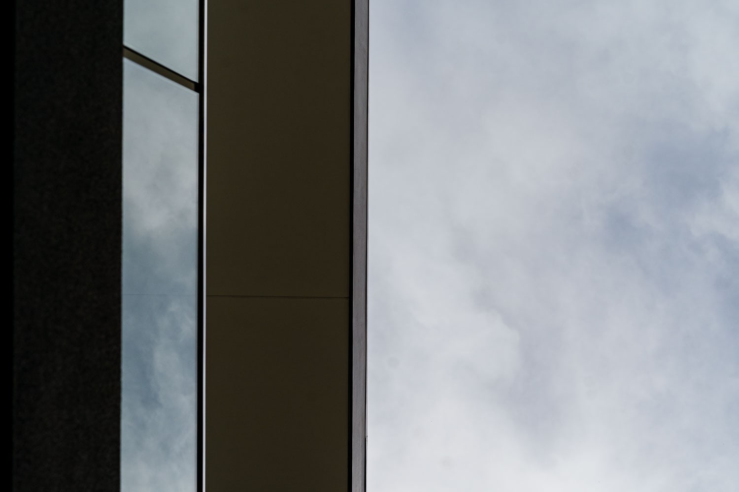 Tight architectural detail of a dark facade corner where a glazed window meets a solid panel, set against an overcast sky.
