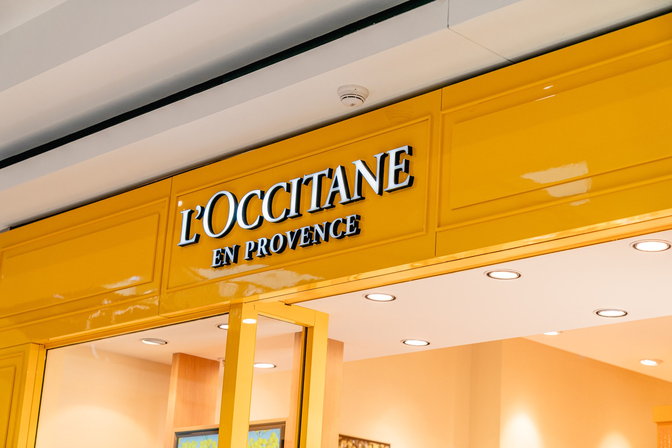 Close-up of the illuminated L'Occitane en Provence storefront sign in glossy yellow above the boutique entrance in a shopping centre.
