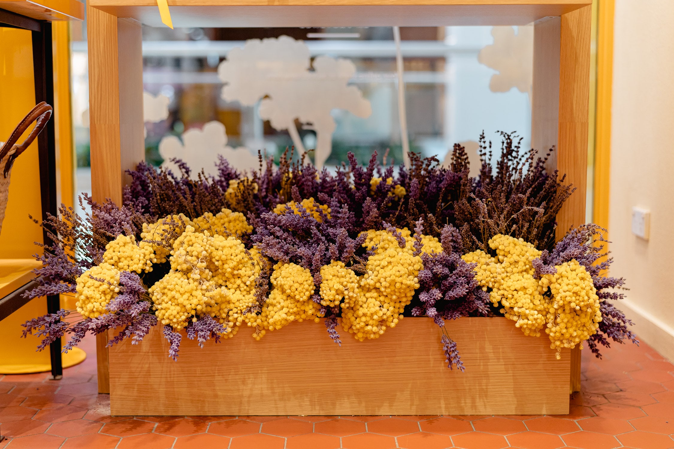 In-store floral display of dried yellow immortelle and purple lavender arranged in a wooden trough box against a Provence-themed window.