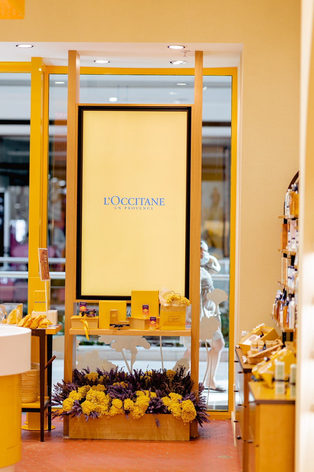 Boutique product display beneath a backlit L'Occitane en Provence lightbox, with skincare boxes above a base of yellow and lavender flowers.