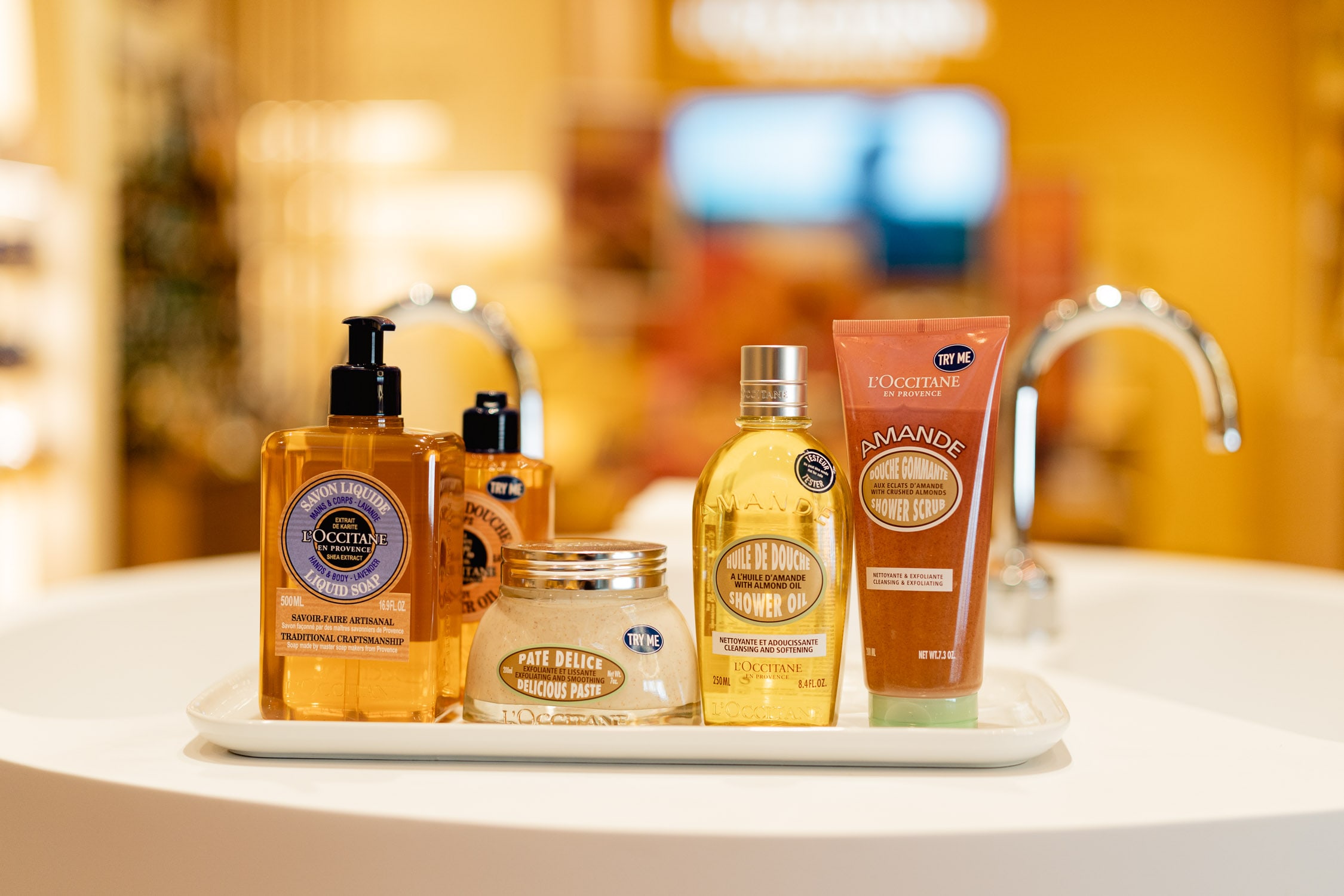L'Occitane shea butter and almond bath products, including liquid soap, shower oil and scrub, arranged on a white tray at a store basin.