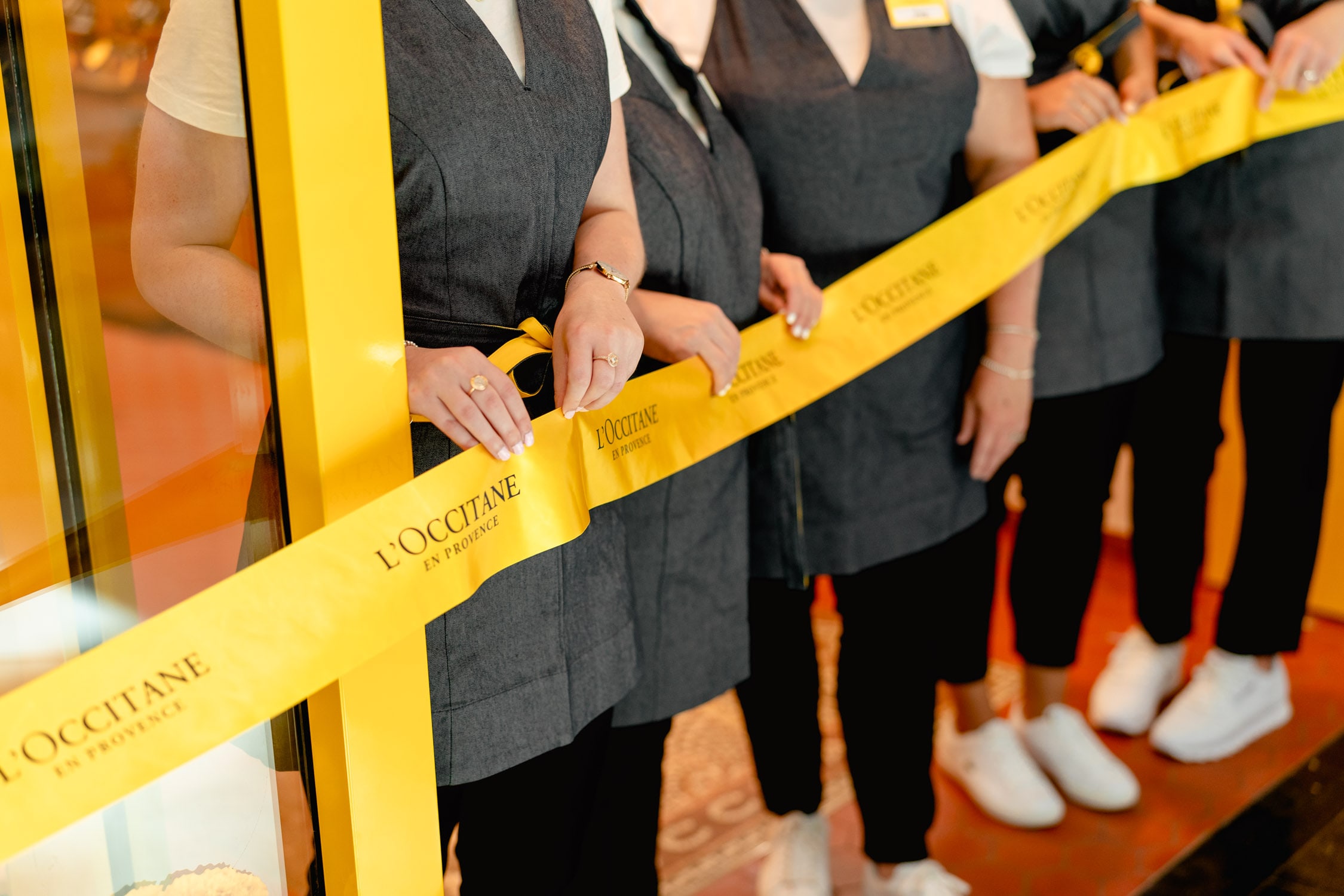 L'Occitane staff in charcoal aprons stand behind a yellow branded ribbon at the store, one holding it for a ribbon-cutting ceremony.