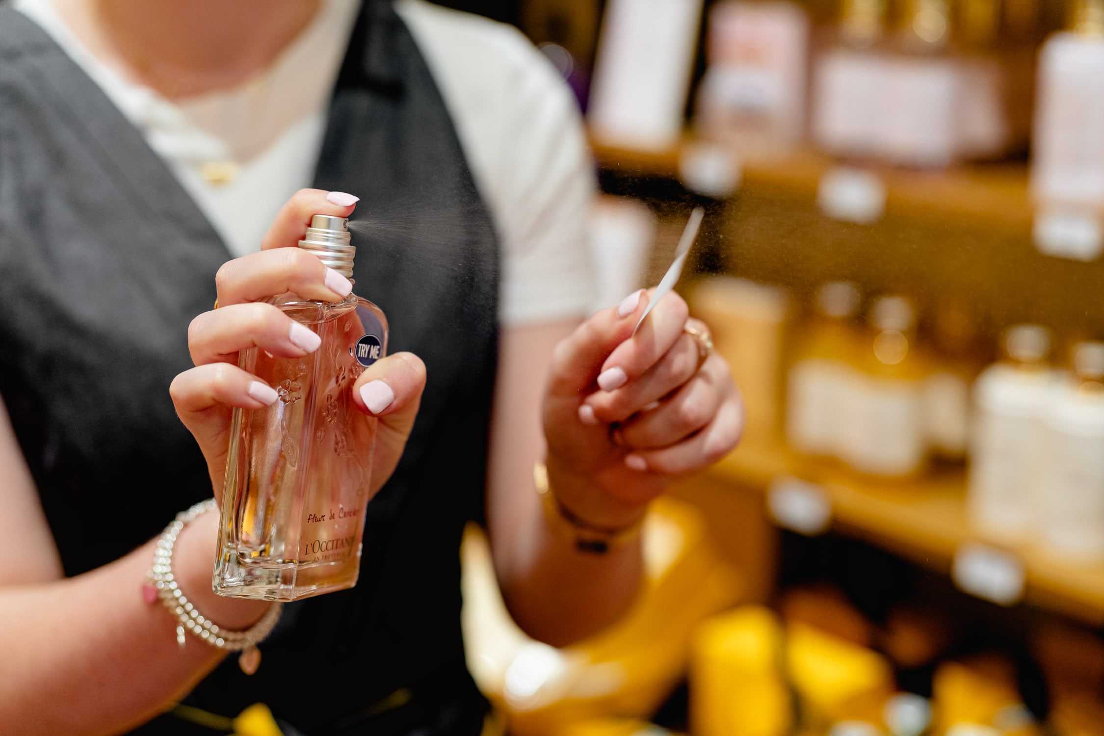 A L'Occitane consultant in an apron sprays Fleur de Cerisier fragrance onto a scent strip amid warmly lit product shelves.