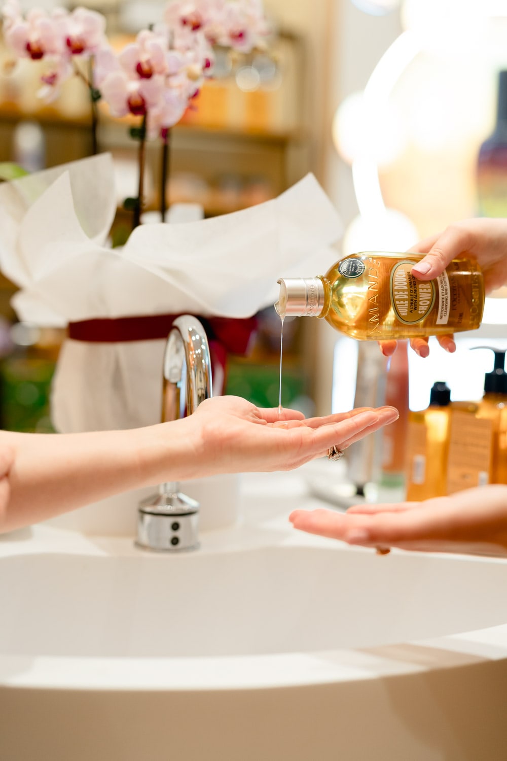 A L'Occitane consultant pours almond shower oil into a guest's cupped hand at a store basin beside a pink orchid and gift-wrapped bouquet.