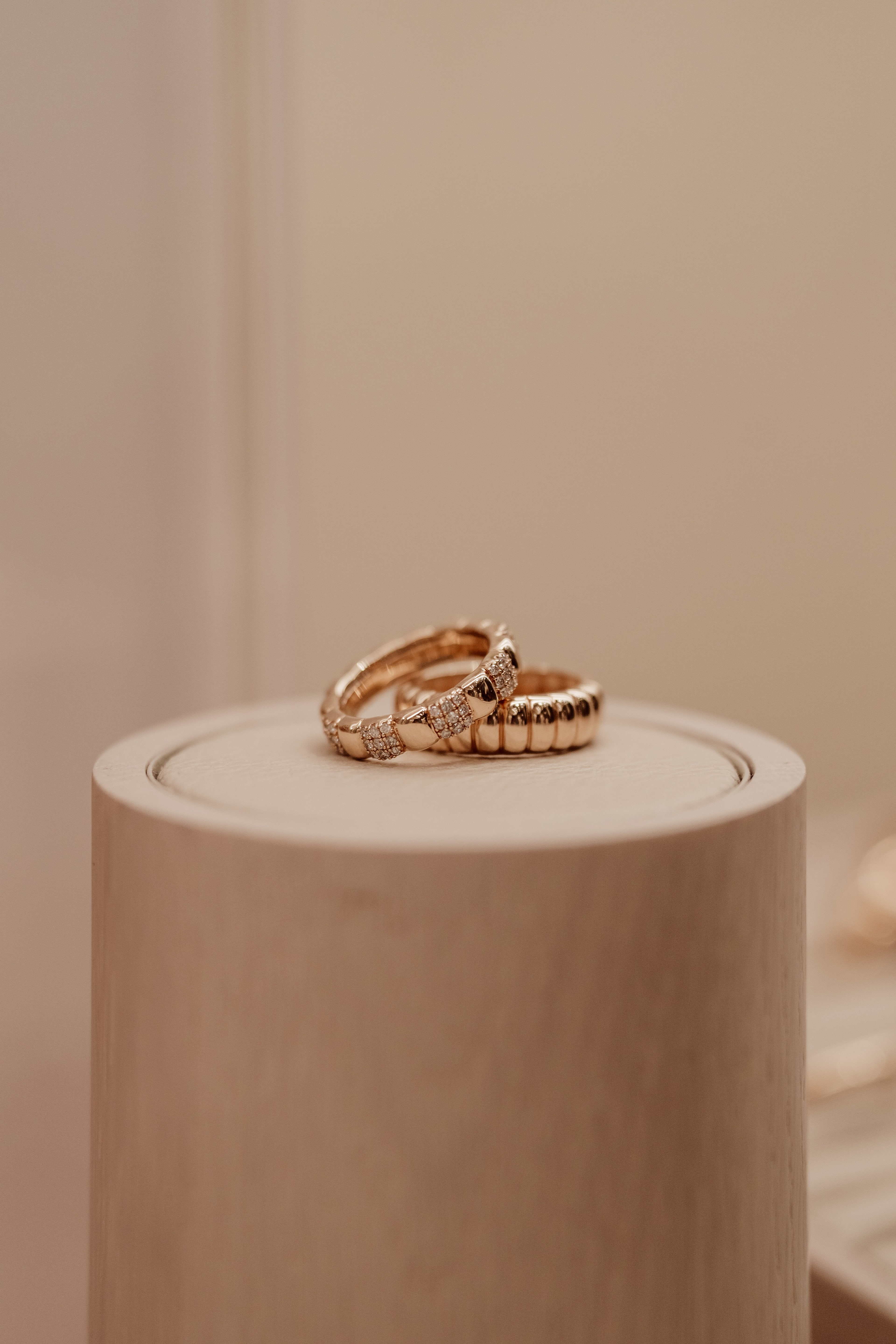 Two gold Mejuri rings, one ridged and one diamond-pave, styled atop a pale wood cylindrical plinth against a soft warm neutral backdrop.