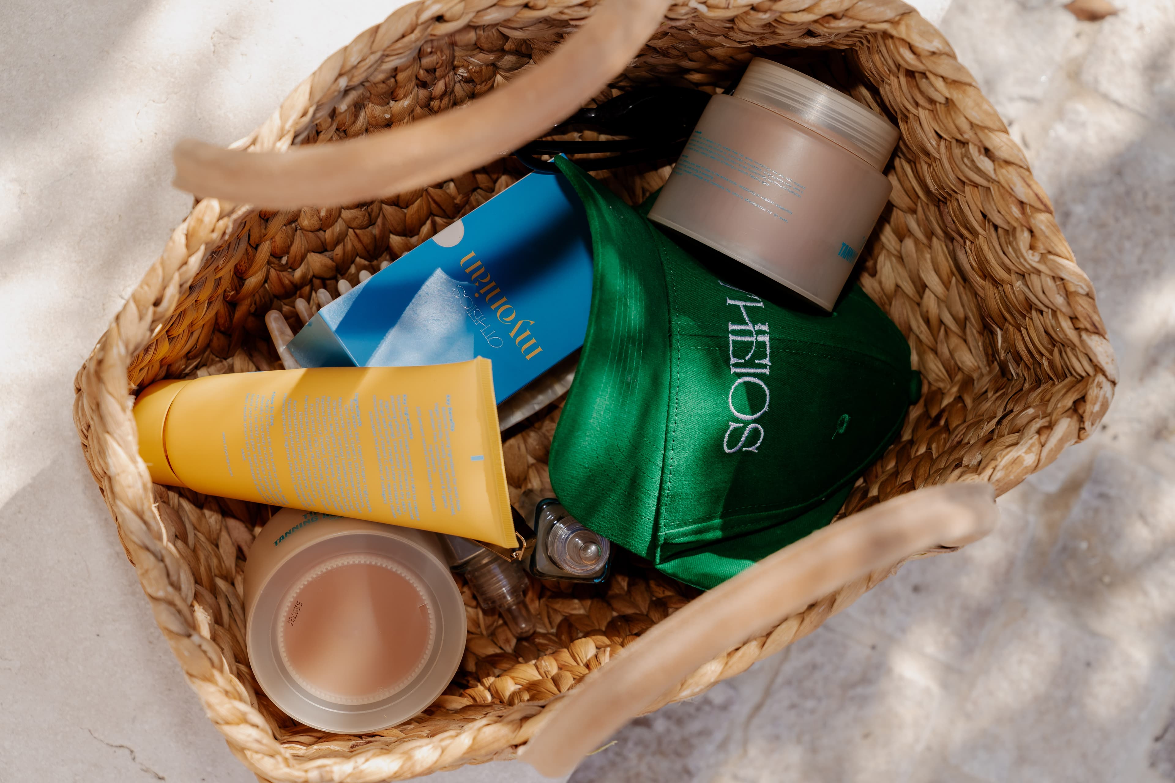 Overhead view of a woven straw basket holding Otheios products, a green branded cap, yellow tube, and jars on a sunlit stone floor.