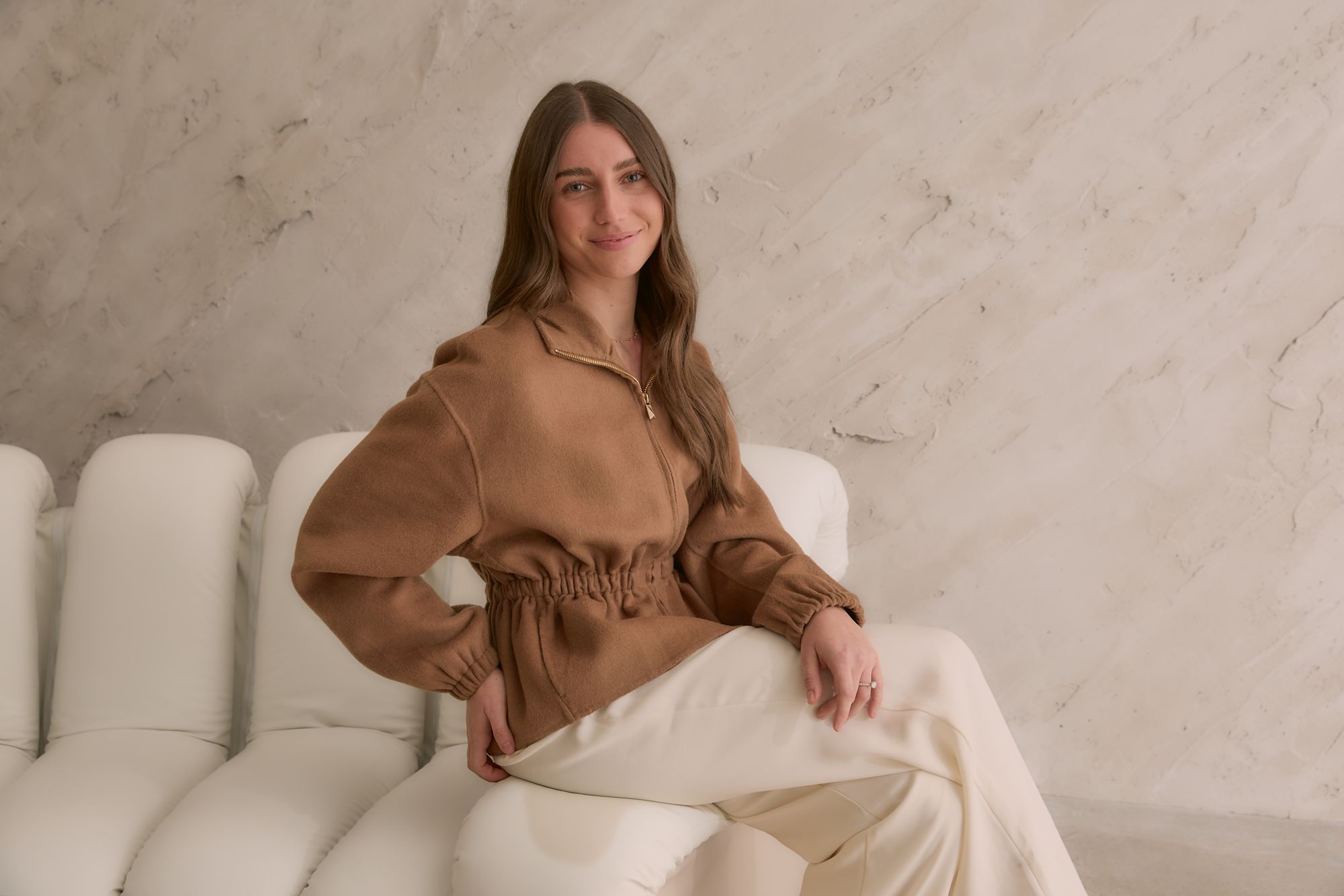 A Peter Petinos Creative team member in a camel jacket seated against a marble studio wall.