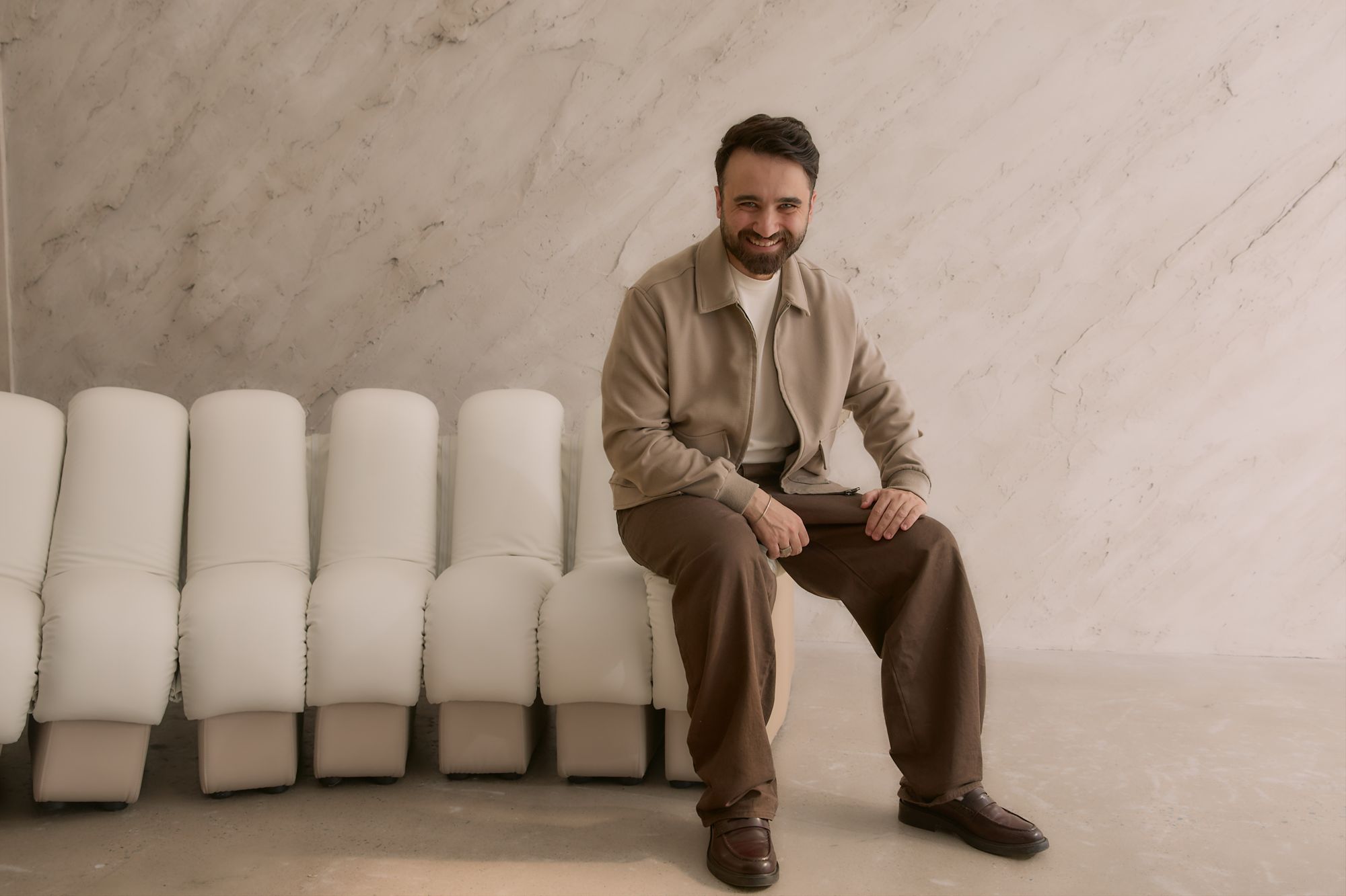 A Peter Petinos Creative team member in a beige jacket seated on studio seating, smiling.