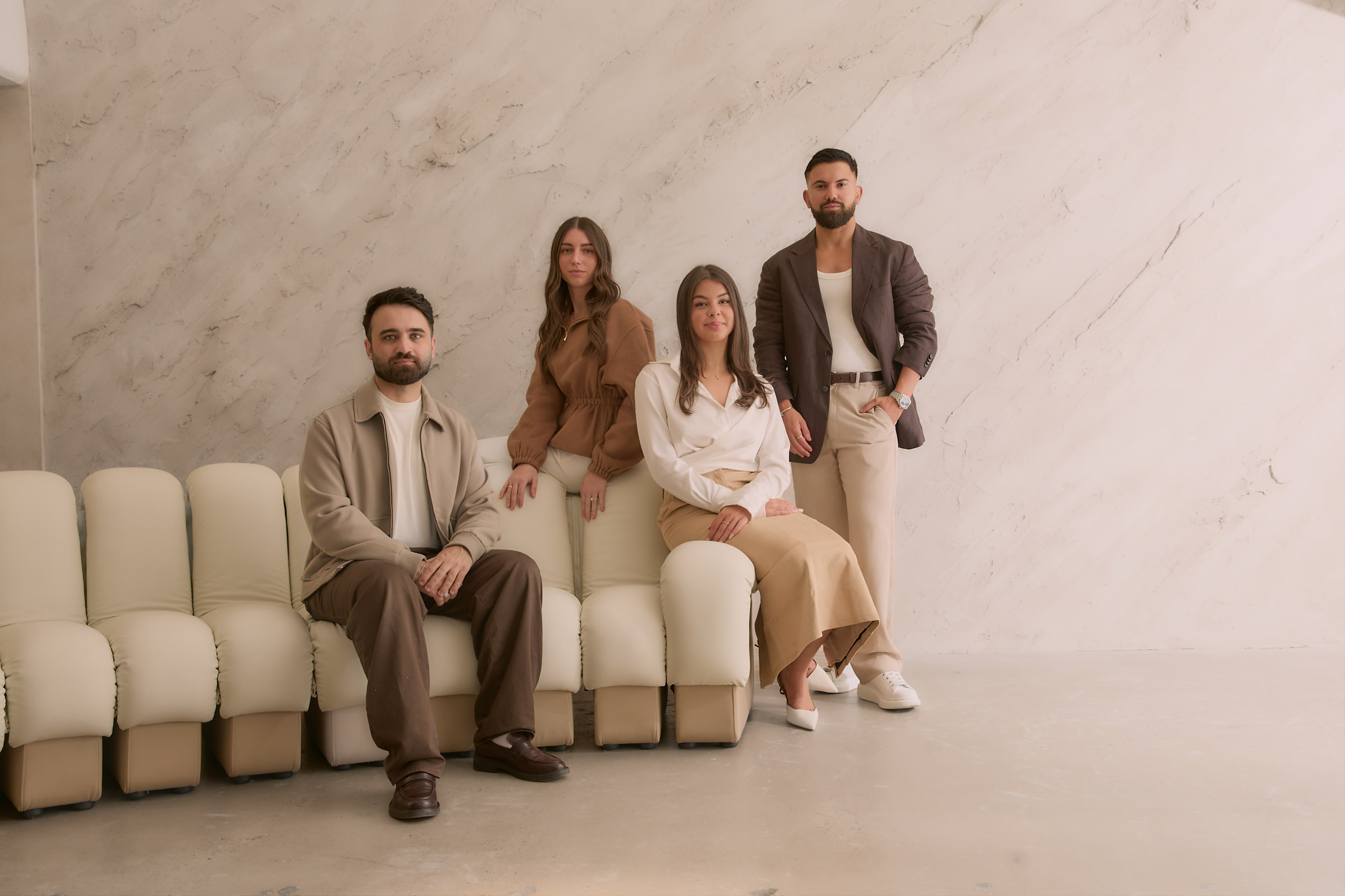 The Peter Petinos Creative team seated together on a row of cream chairs against a marble wall.