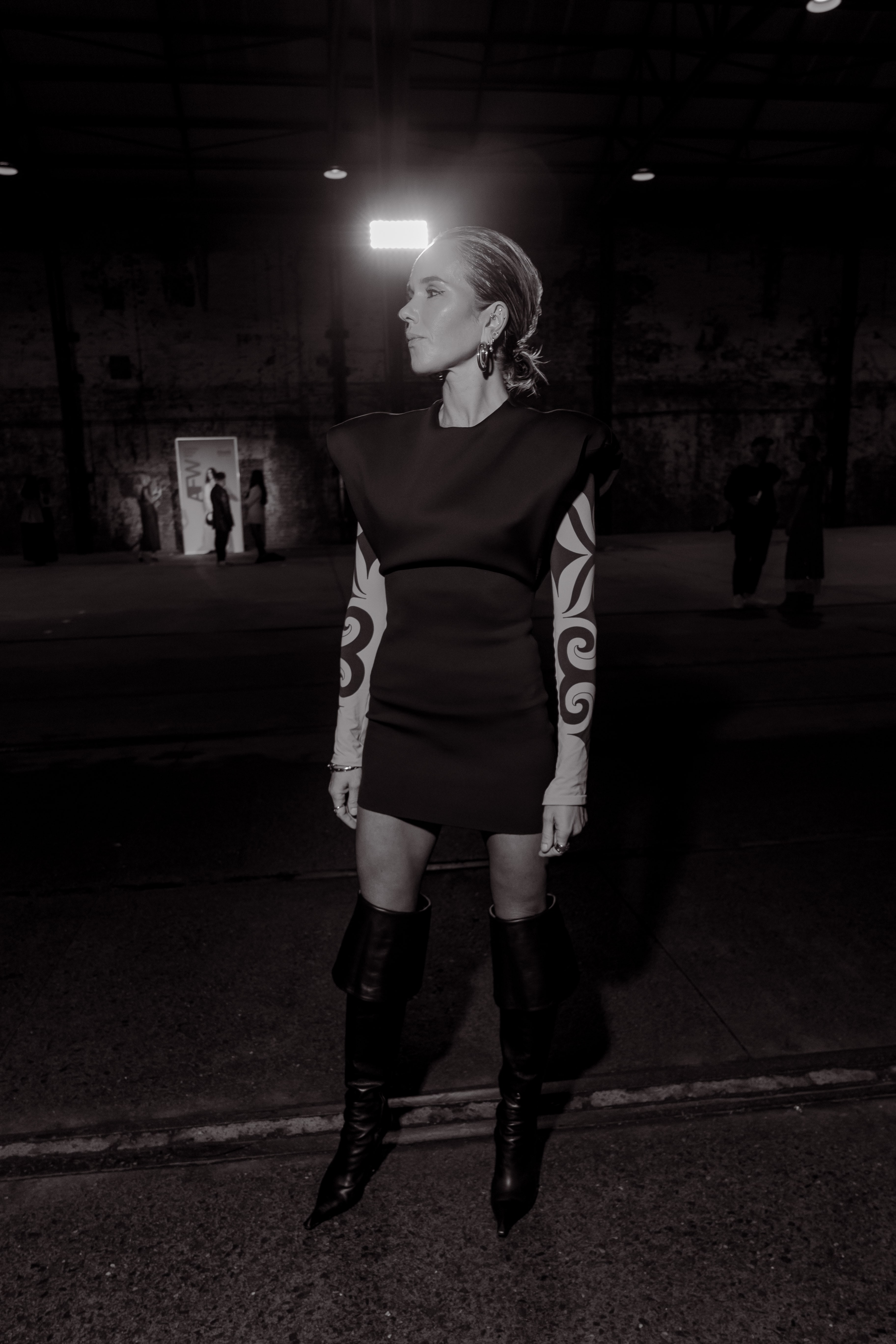 Black-and-white shot of a woman in a structured mini dress with swirl-patterned sleeves and knee-high boots under a spotlight in a warehouse.