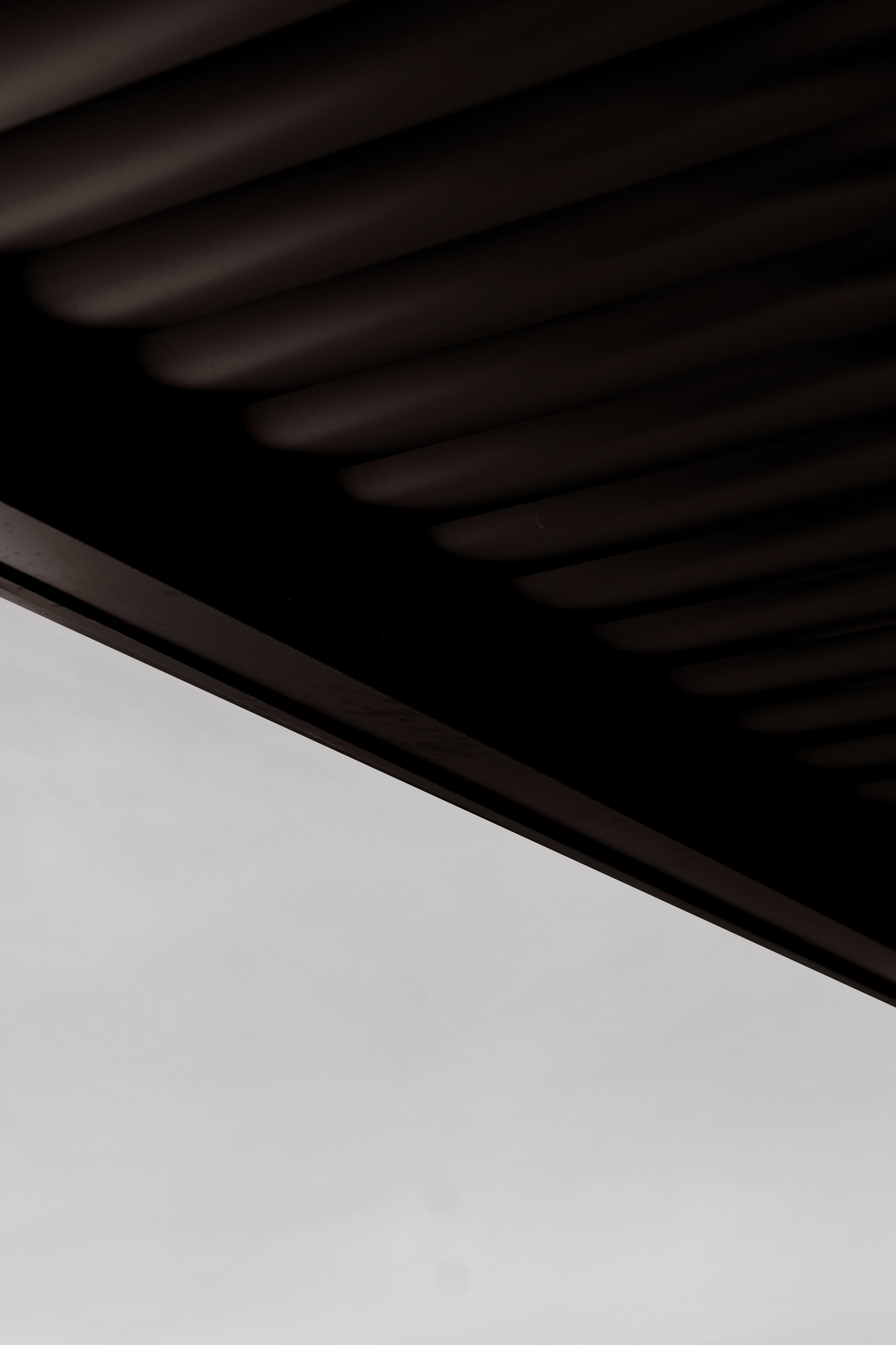 Underside of a dark ribbed timber-batten soffit or ceiling cutting a diagonal across a pale grey sky.