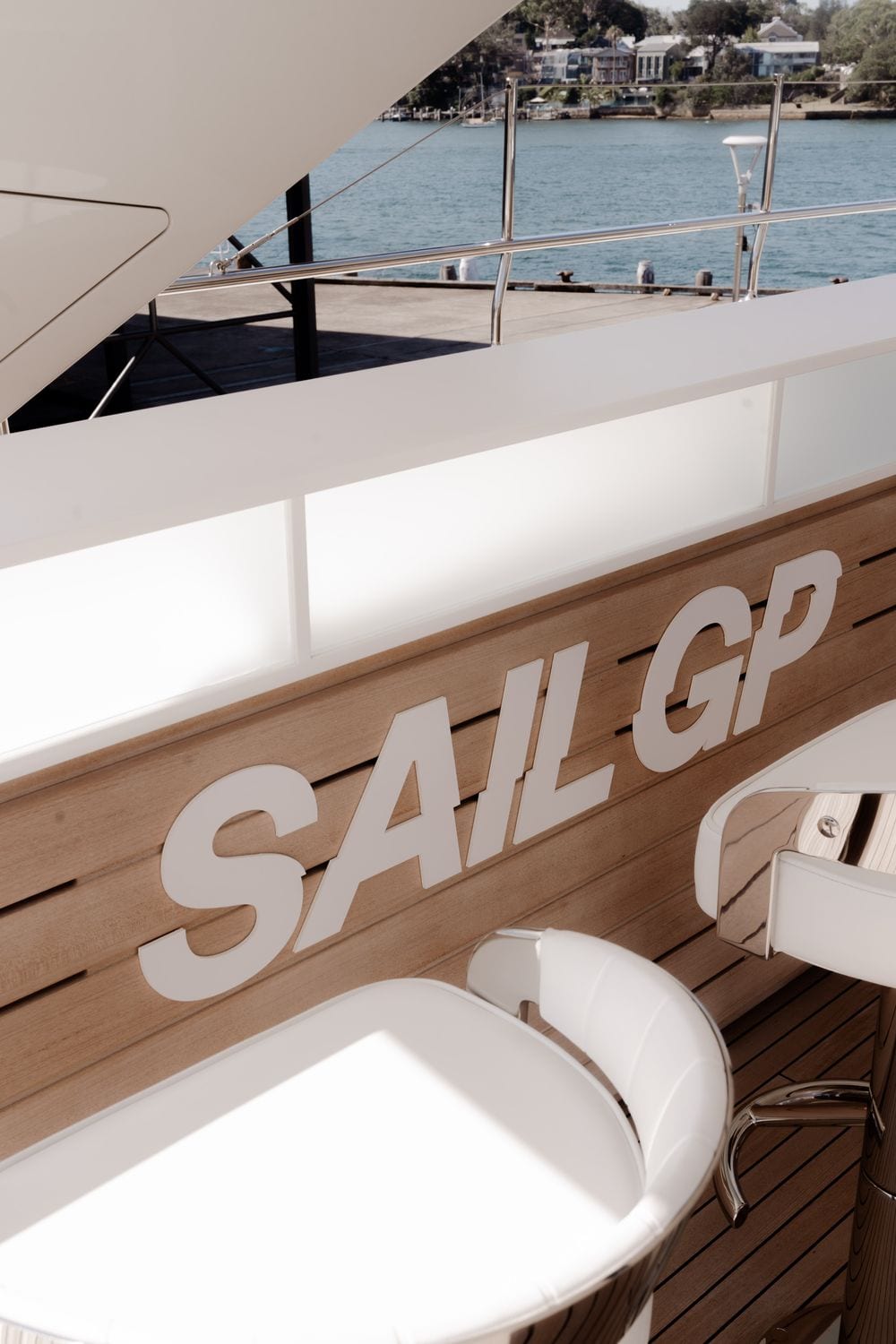 Superyacht deck detail with 'SAIL GP' branding on teak panelling, a white bar stool, and Sydney Harbour visible beyond the railing.