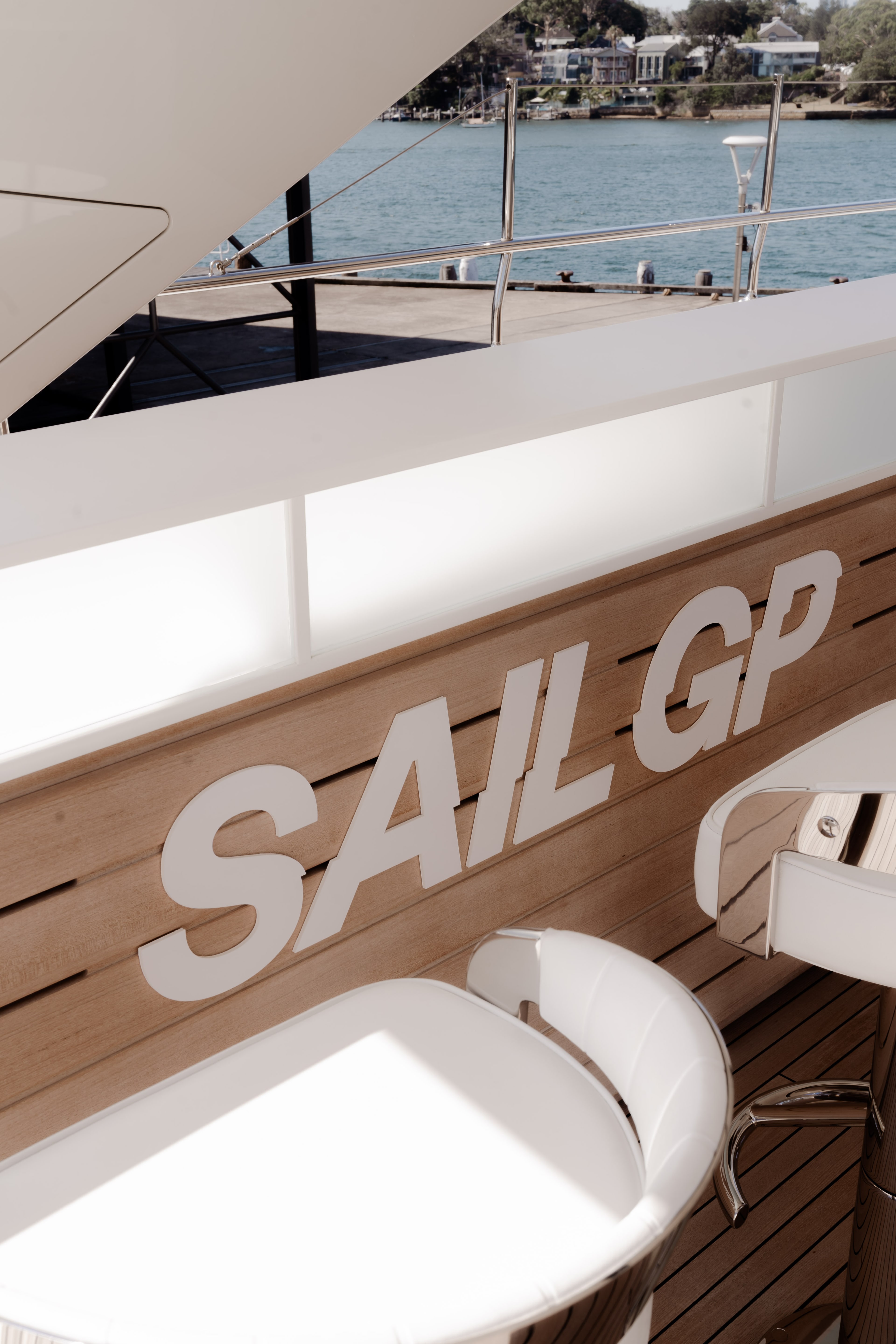 Superyacht deck detail with 'SAIL GP' branding on teak panelling, a white bar stool, and Sydney Harbour visible beyond the railing.