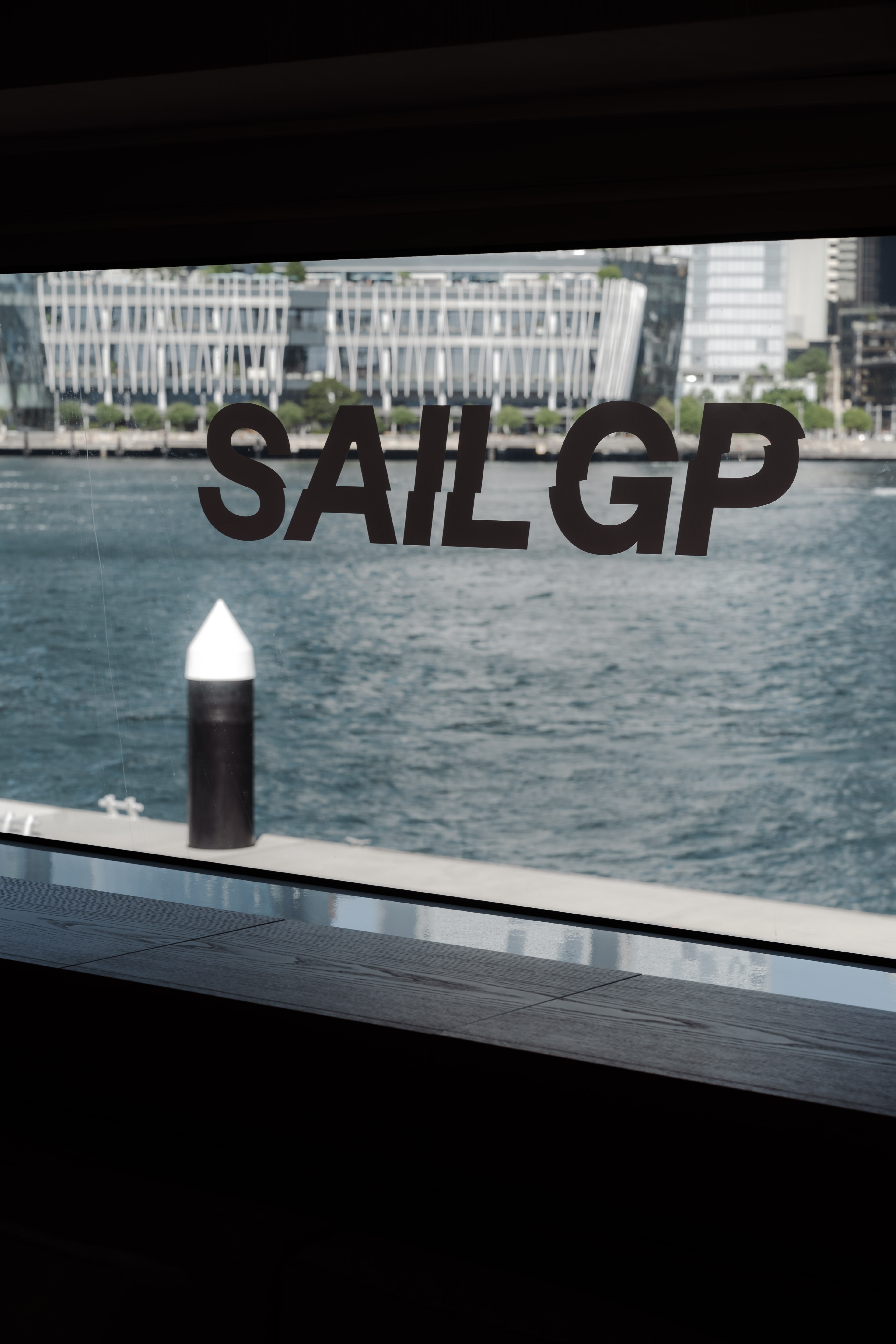 View through a yacht window bearing the 'SAIL GP' logo, looking across Sydney Harbour to a mooring pylon and waterfront buildings.