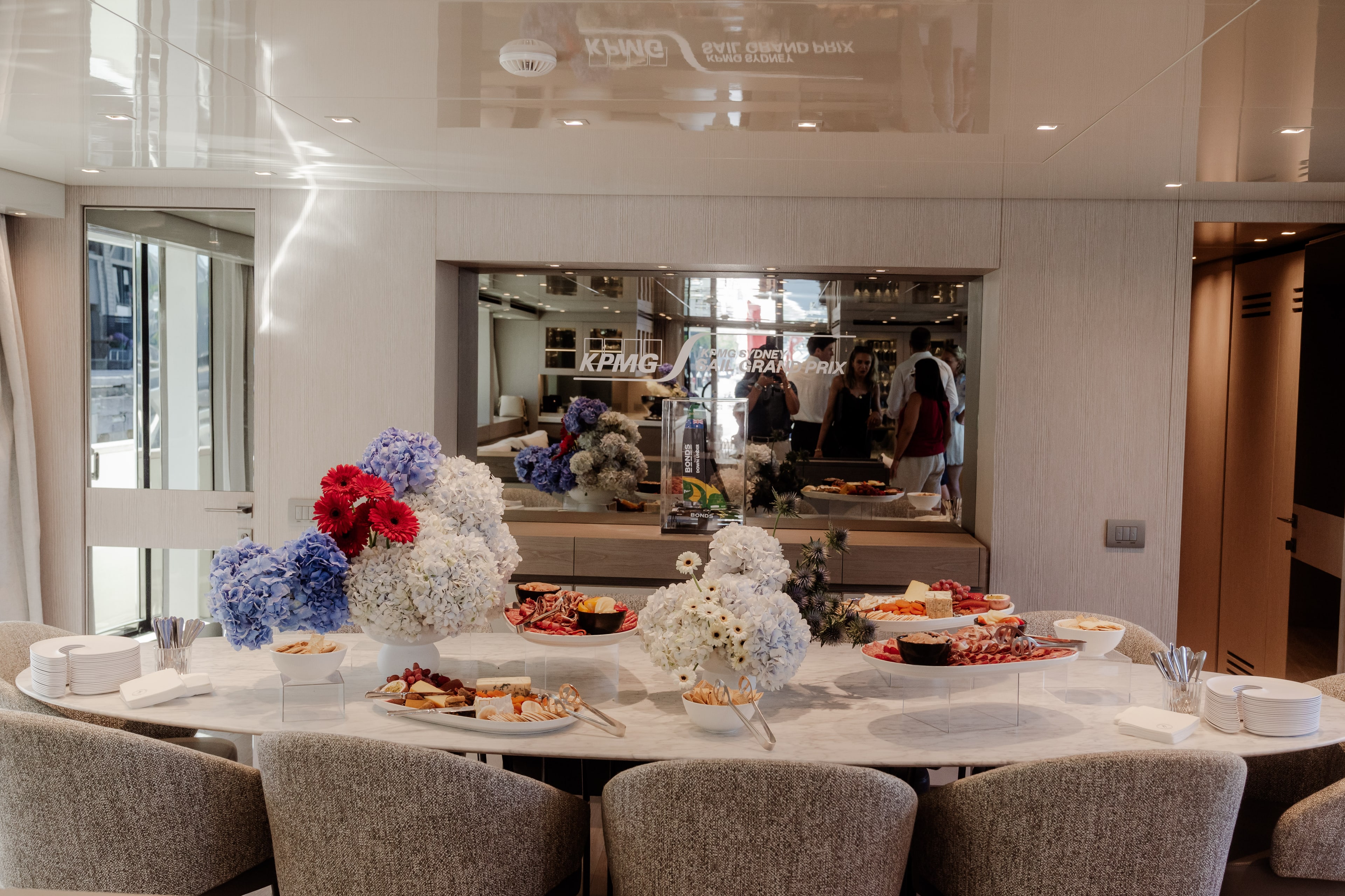 Yacht dining table laid with canapes and blue, white and red hydrangea arrangements, with KPMG SailGP branding and guests behind glass.
