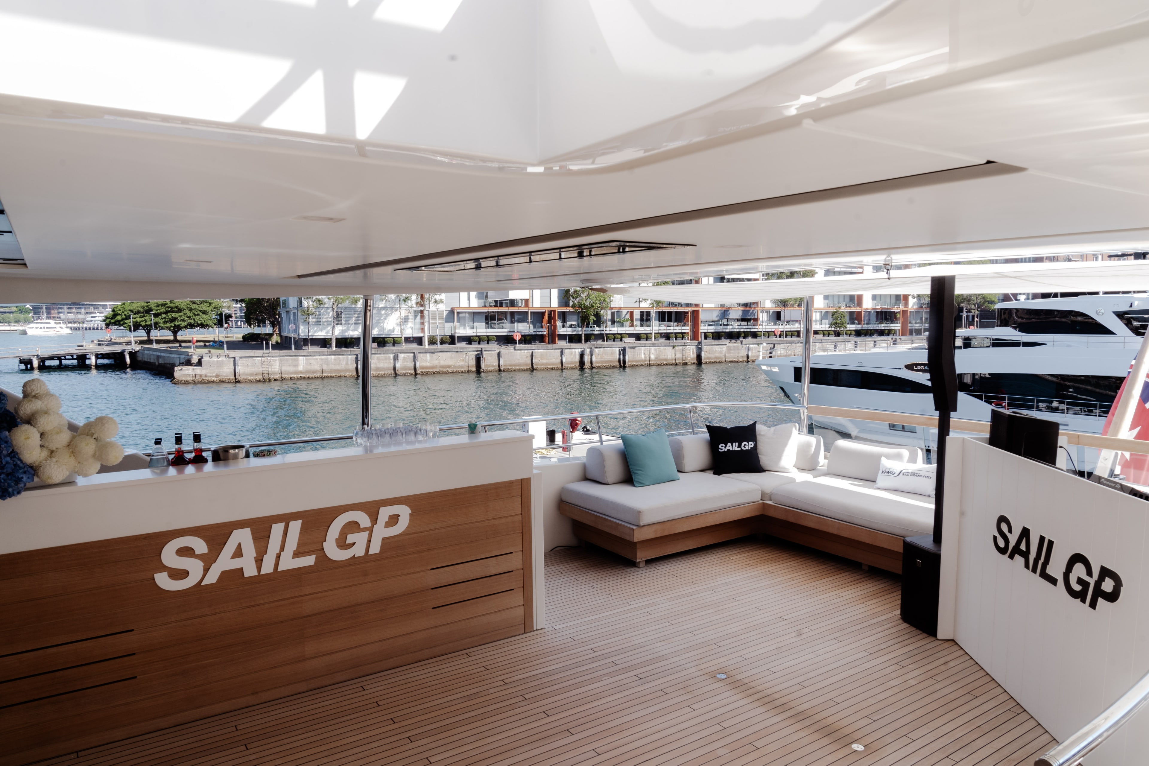 Sunlit superyacht aft deck with 'SAIL GP' branded panels, cushioned lounge seating and logo cushions, overlooking a Sydney Harbour marina.