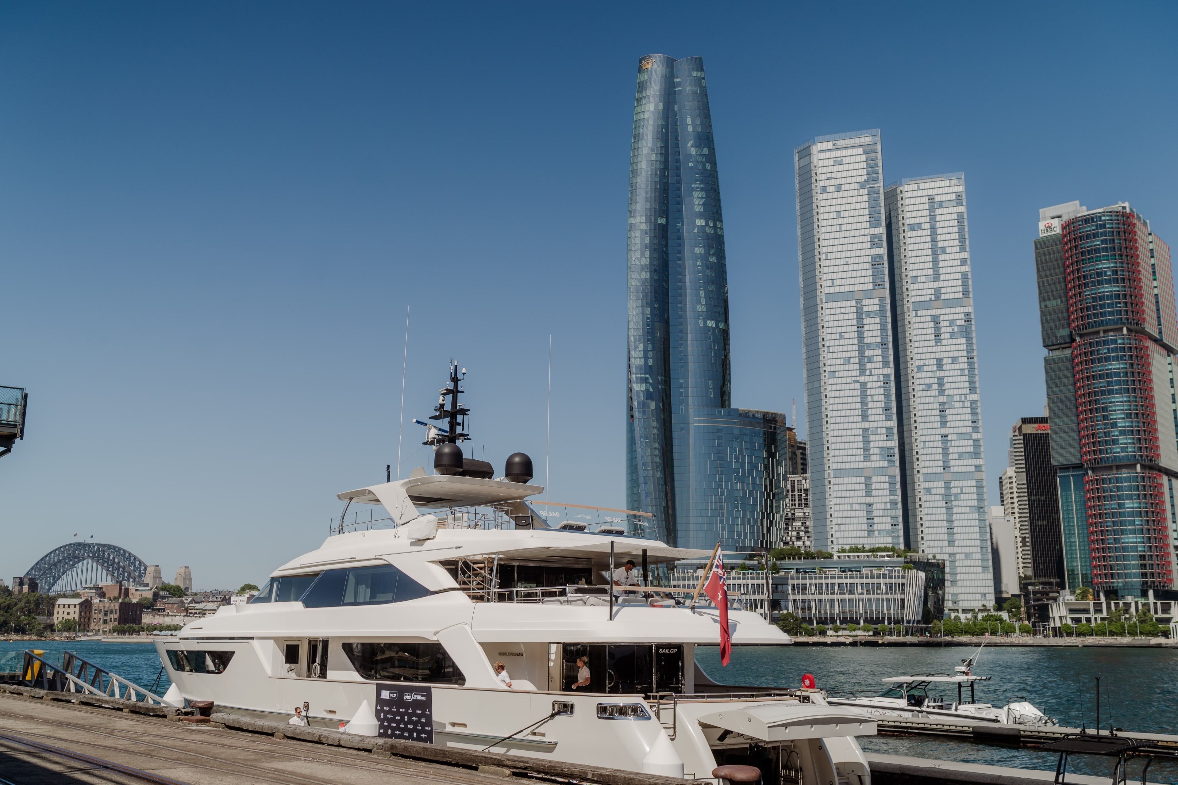 Superyacht LANCE berthed on Sydney Harbour against city skyscrapers, with the Harbour Bridge visible in the background under clear blue sky.