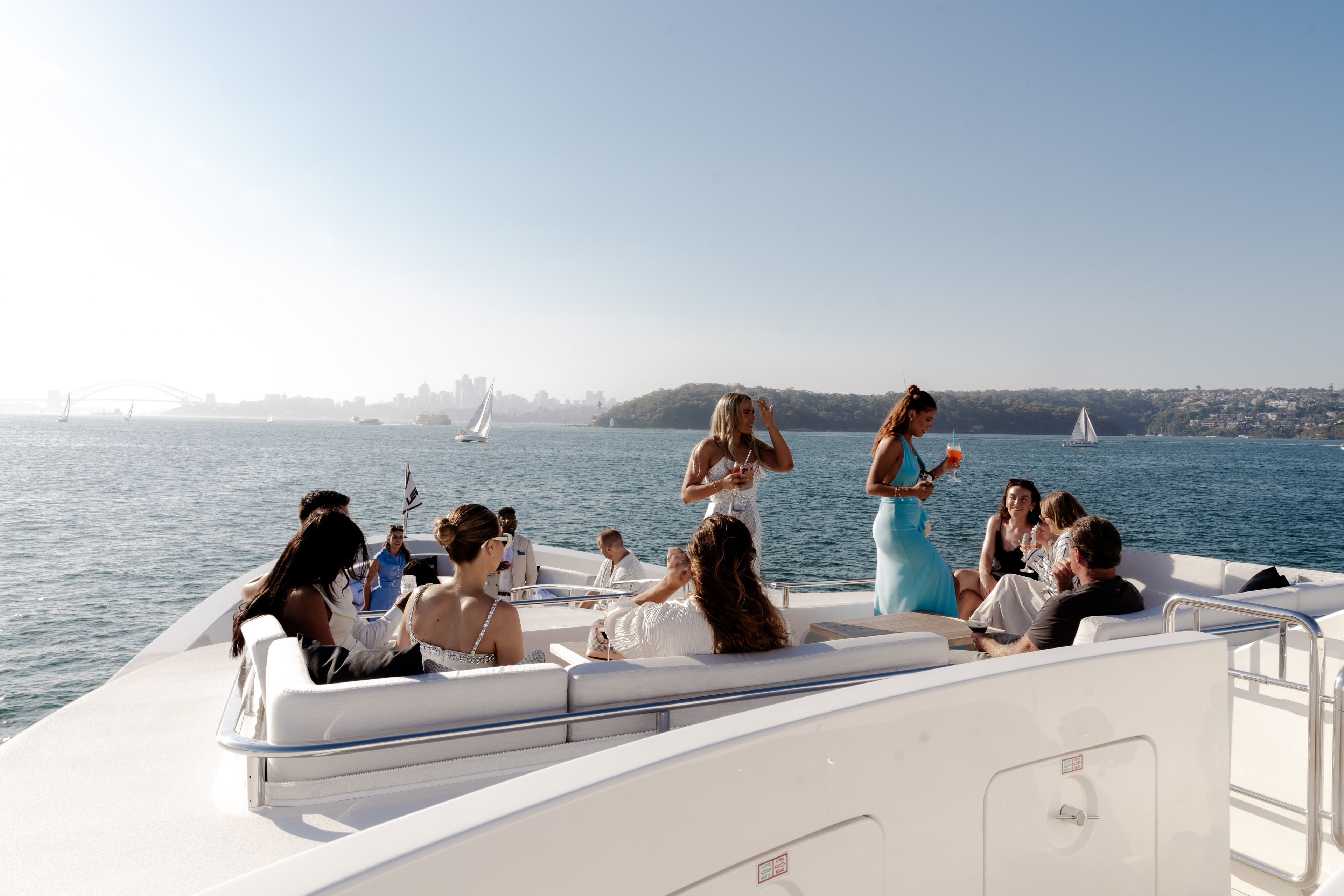 Guests mingle with cocktails on a superyacht foredeck lounge on Sydney Harbour, with sailboats and a hazy headland in the background.