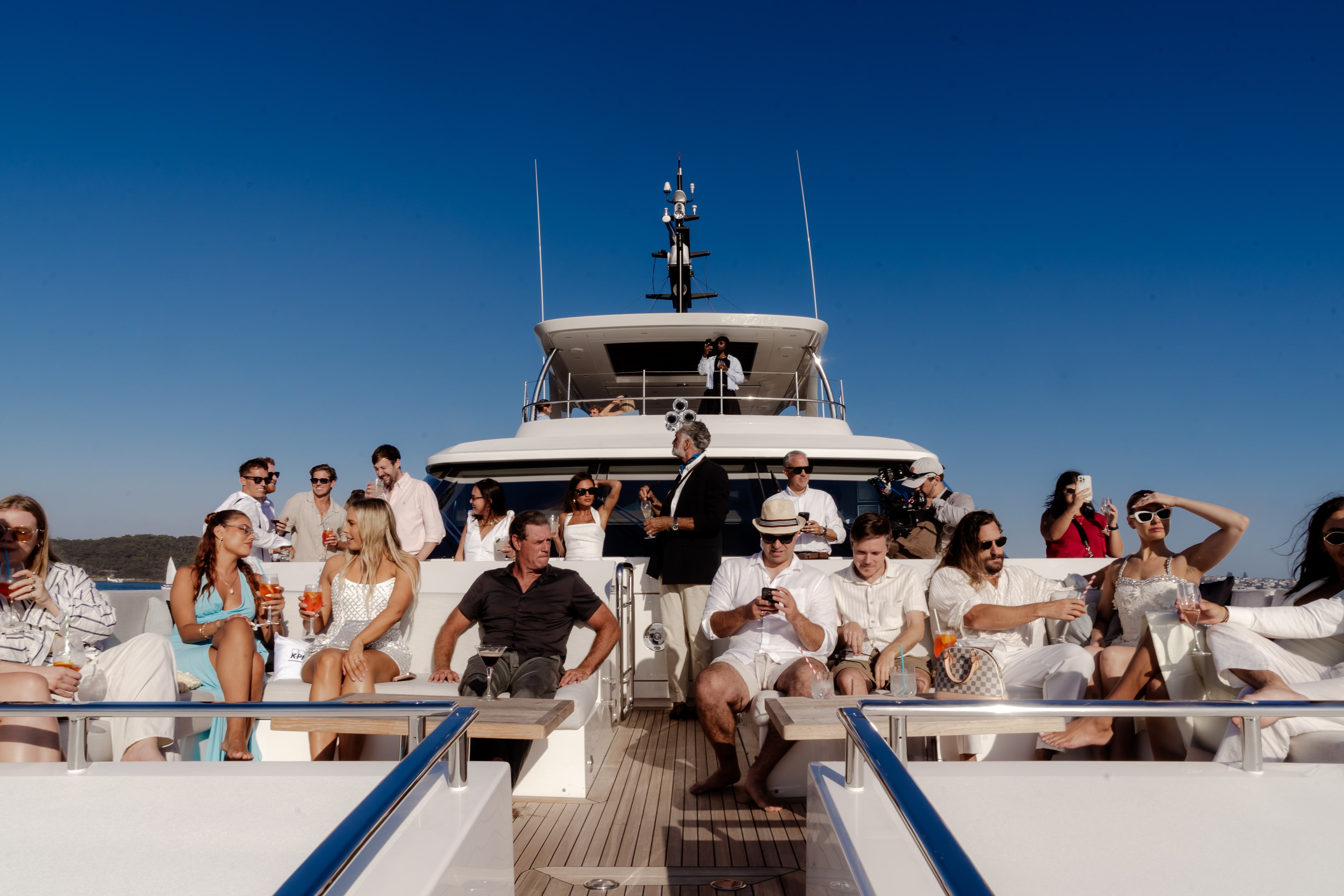 Well-dressed guests relax with drinks on a superyacht deck under bright blue sky on Sydney Harbour during the SailGP event.